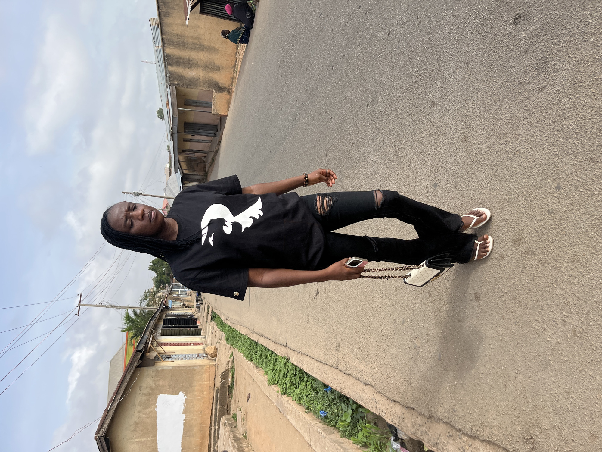 A medium shot captures a person in Jos, Nigeria, standing on an asphalt road. The individual, with dark skin, is wearing a black T-shirt with a white graphic design on the front, ripped black jeans, and white flip-flops. A beaded bracelet is visible on the person's wrist. They are holding a white purse with a gold chain strap.
The background shows a neighborhood with buildings of varying heights, primarily beige in color. Some buildings have corrugated metal roofs. A narrow green strip of vegetation runs along the side of the road. Overhead power lines are visible against a cloudy sky.
