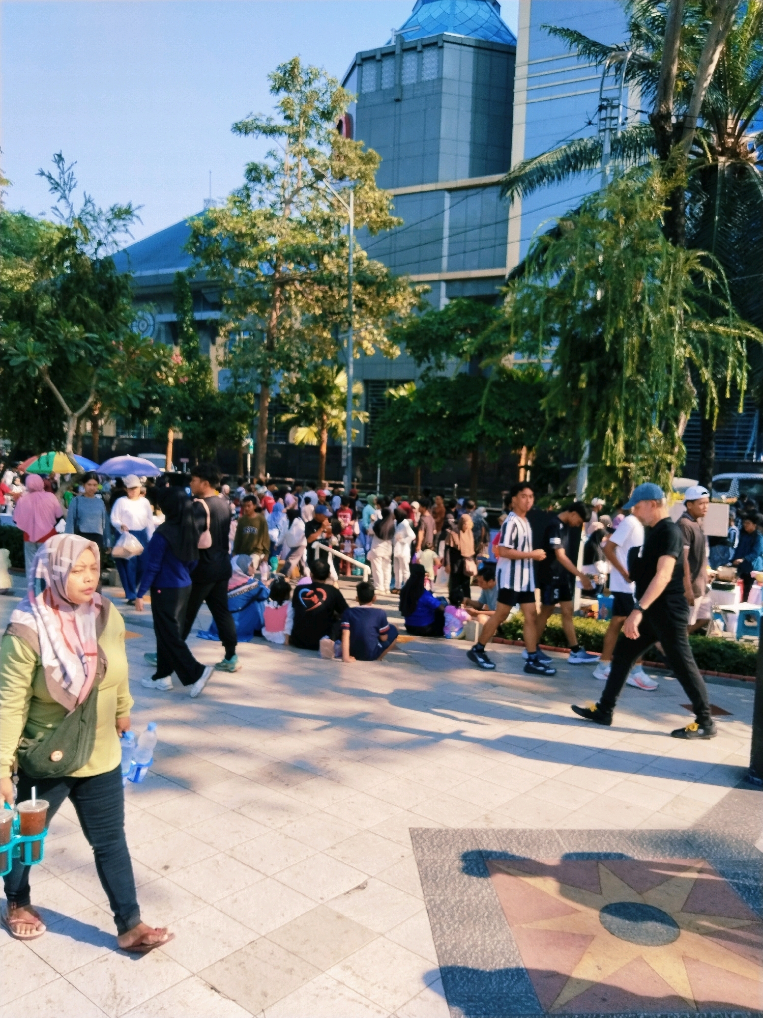 The image depicts a public gathering in Surabaya, Indonesia, likely at an outdoor venue adjacent to modern buildings. In the foreground, a woman wearing a light green long-sleeved shirt and dark jeans is walking, carrying a drink carrier with two iced beverages and a bottle of water. She has a beige hijab with pink and white patterns and a green shoulder bag. The ground is paved with square tiles, and some have decorative patterns. A large shadow of a sun-like emblem is cast on the pavement.
In the midground and background, a crowd of people is milling about. Many are dressed casually, some in sportswear. There are trees and foliage scattered throughout the scene, with lush green leaves. In the distance, a tall, modern building with blue-tinted windows and a domed blue roof dominates the skyline, suggesting an urban environment. Some individuals are seated on the ground, while others are standing or walking. There are also some makeshift stalls or vendors visible, indicated by umbrellas and various items displayed. The lighting suggests it is daytime, possibly late afternoon, with long shadows cast on the ground. The overall atmosphere appears to be one of a community event or casual social gathering.