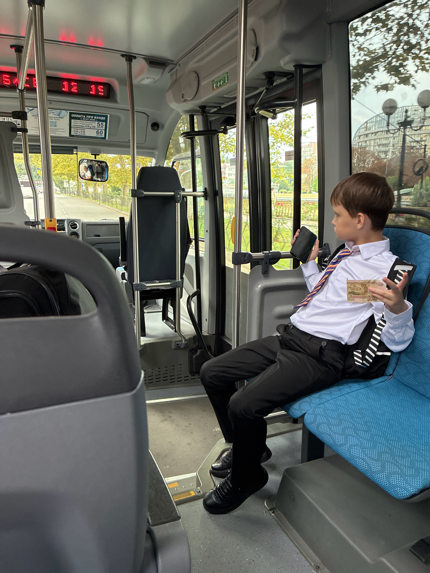 A young boy, dressed in a white dress shirt, black tie, and black pants, sits on a blue bus seat in Sochi, Russia. He holds a black smartphone in his left hand and a 100 ruble note in his right hand, appearing to be either showing it or preparing to pay. The bus interior is visible, with metal poles, a driver's compartment in the distance, and windows showing a street scene with trees and buildings. The digital display above the driver's area shows "01 15" in red LEDs. A sign detailing bus fares is also visible. The boy has short brown hair and is looking to his right.