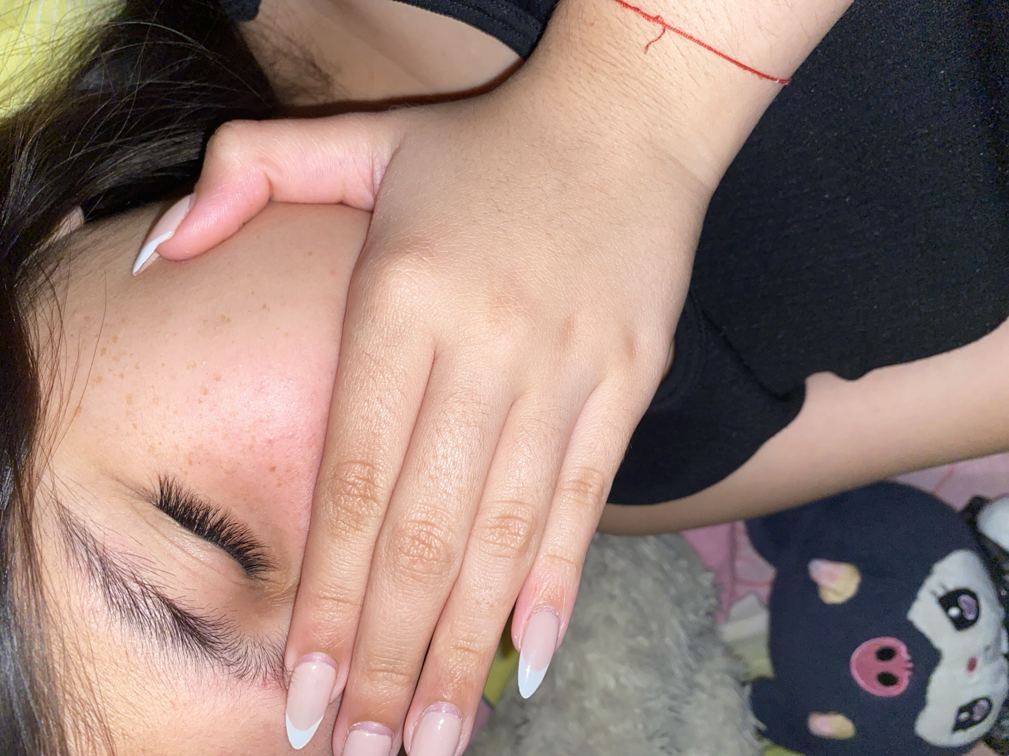 A close-up, indoor shot shows a person's face partially obscured by their hand. The person has dark hair, light skin with freckles, and long eyelashes. Their eyes are closed. The hand covering their face has light skin and long, pointed, French-manicured nails. A red string is visible on the wrist. The person is wearing a black top. In the background, there is a plush toy, dark blue with white and pink details, sitting on what appears to be a bed with pink and yellow patterned sheets. Cotoperíz, Venezuela is not apparent in this image.