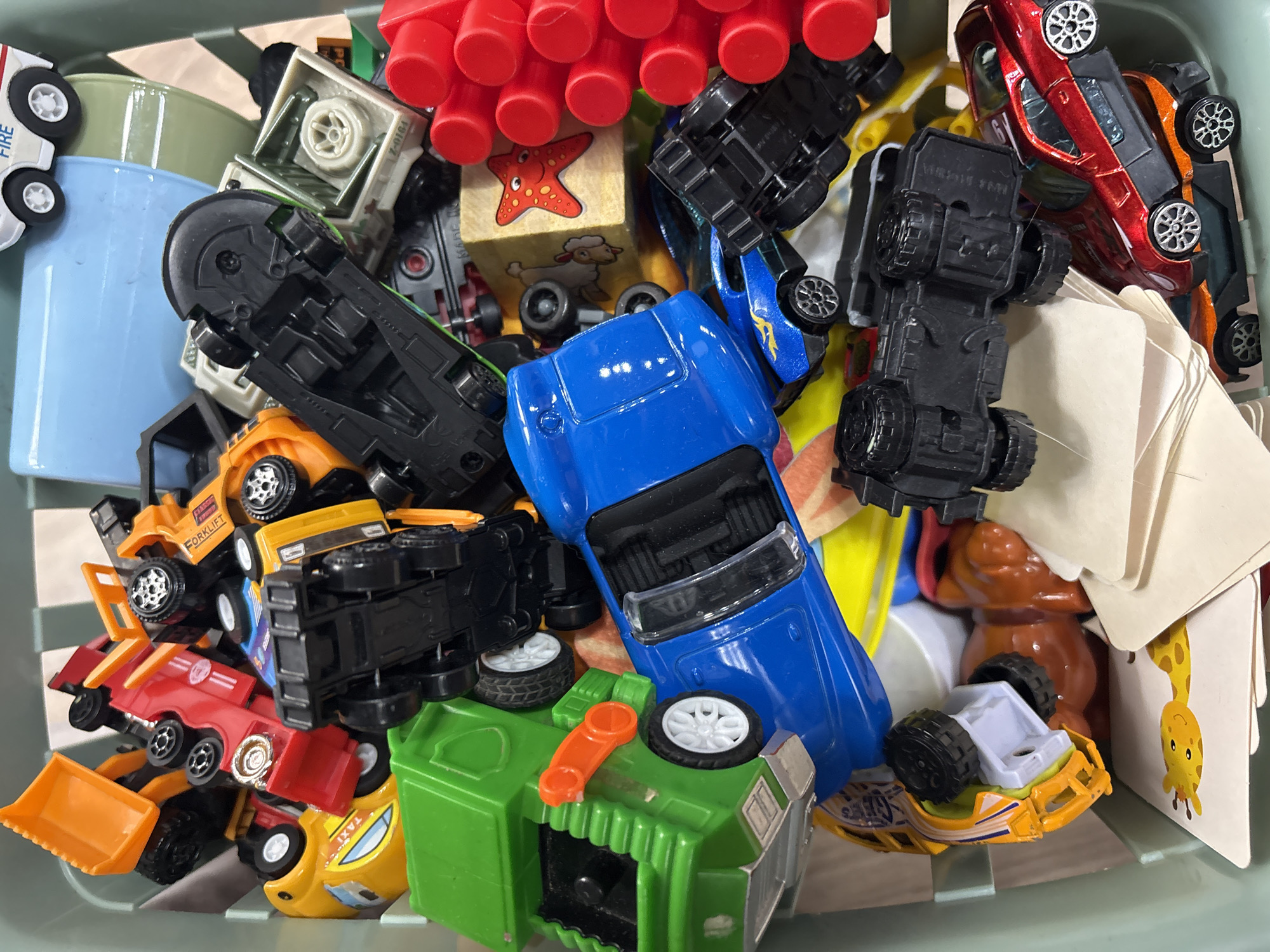 The image shows a green plastic bin filled with various toy vehicles. Among the toys are multiple cars, a dump truck, a garbage truck, and a forklift. The vehicles are predominantly made of plastic and metal, with a variety of colors including red, blue, yellow, and green. Some of the vehicles are upside down, and others are stacked on top of each other. In the background, a blue plastic container is partially visible. The toys appear to be well-used. The environment is consistent with a toy box or storage bin in a home. There is no indication of the specific location within Mikhaylovsk, Russia, as the image only contains toys.