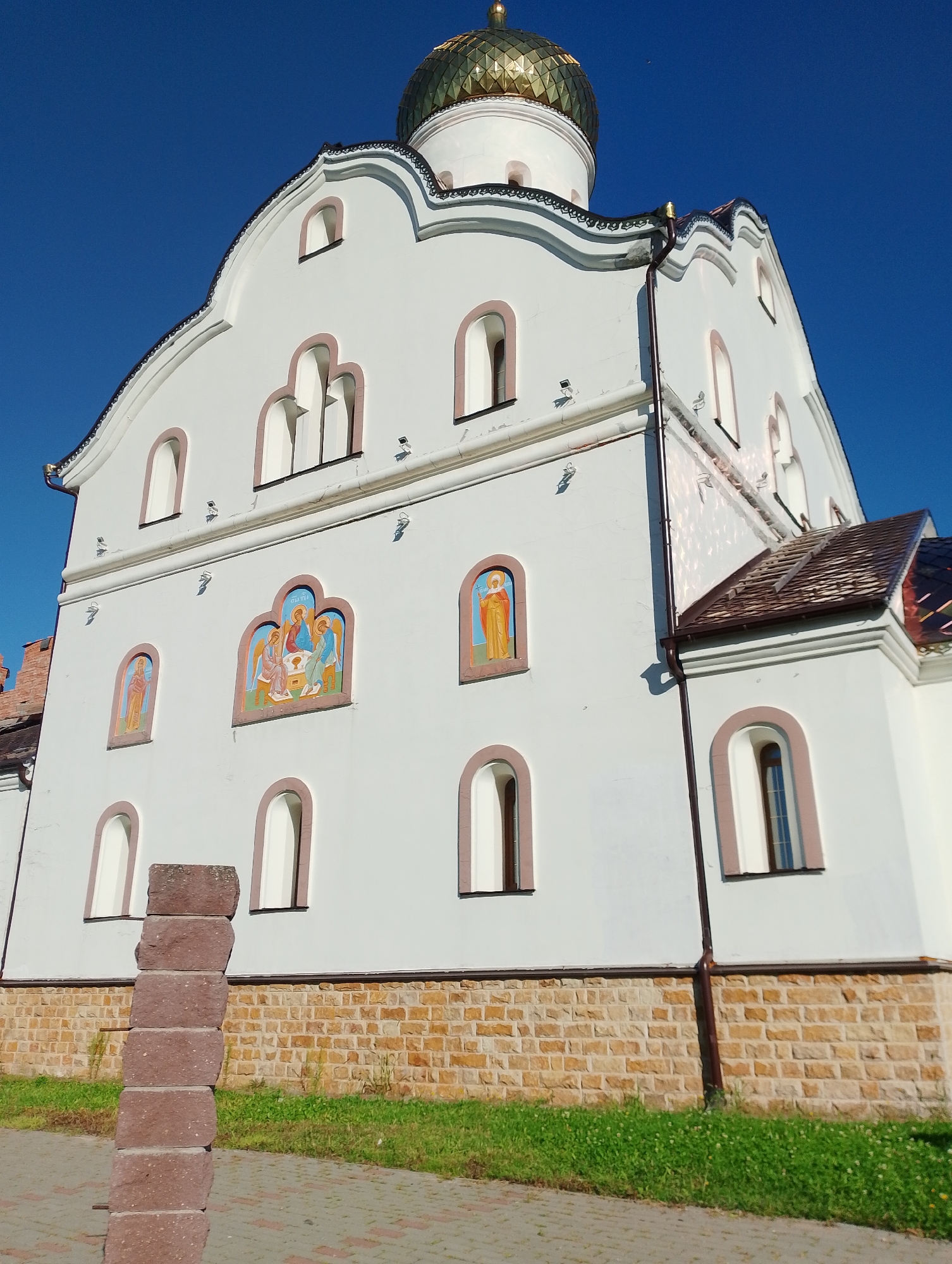 This is a low-angle, eye-level, outdoor, full shot of a white church with a golden dome, seen against a clear blue sky. The church is located in Vitsebsk, Belarus. The building features arched windows with reddish-brown trim. There are several religious icons painted on the white facade, depicting figures in colorful robes. The lower portion of the church is clad in rough-hewn, tan-colored stone blocks. A stack of reddish-brown stone blocks stands in the foreground, to the left of the frame, partially obscuring the church. Green grass grows along the base of the church and a paved walkway is visible in the foreground. The architecture suggests an Orthodox Christian church.