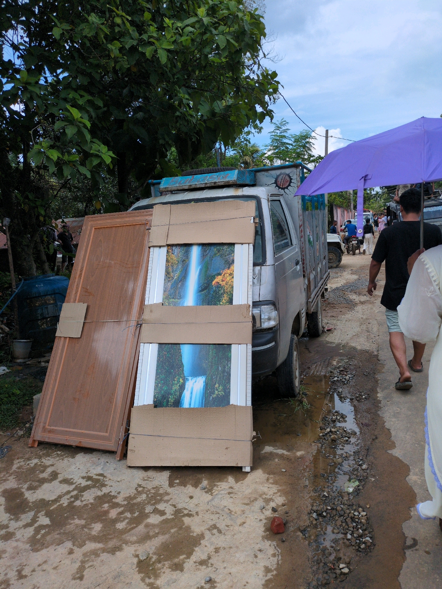 An outdoor scene in Borchora, India, depicts a rural road with a light-colored truck parked on the right side, facing forward. The truck's bed is laden with two large doors, one brown and leaning against the truck, and another with a waterfall image, leaning beside it. A person wearing a black shirt and shorts walks away from the camera, holding a purple umbrella. A woman in a white garment with blue trim walks towards the camera on the right side of the frame. The road is unpaved, with patches of mud and gravel, and a small puddle of water is visible in the foreground. Trees with dense green foliage are visible on the left side of the image. The sky is overcast with some blue visible.