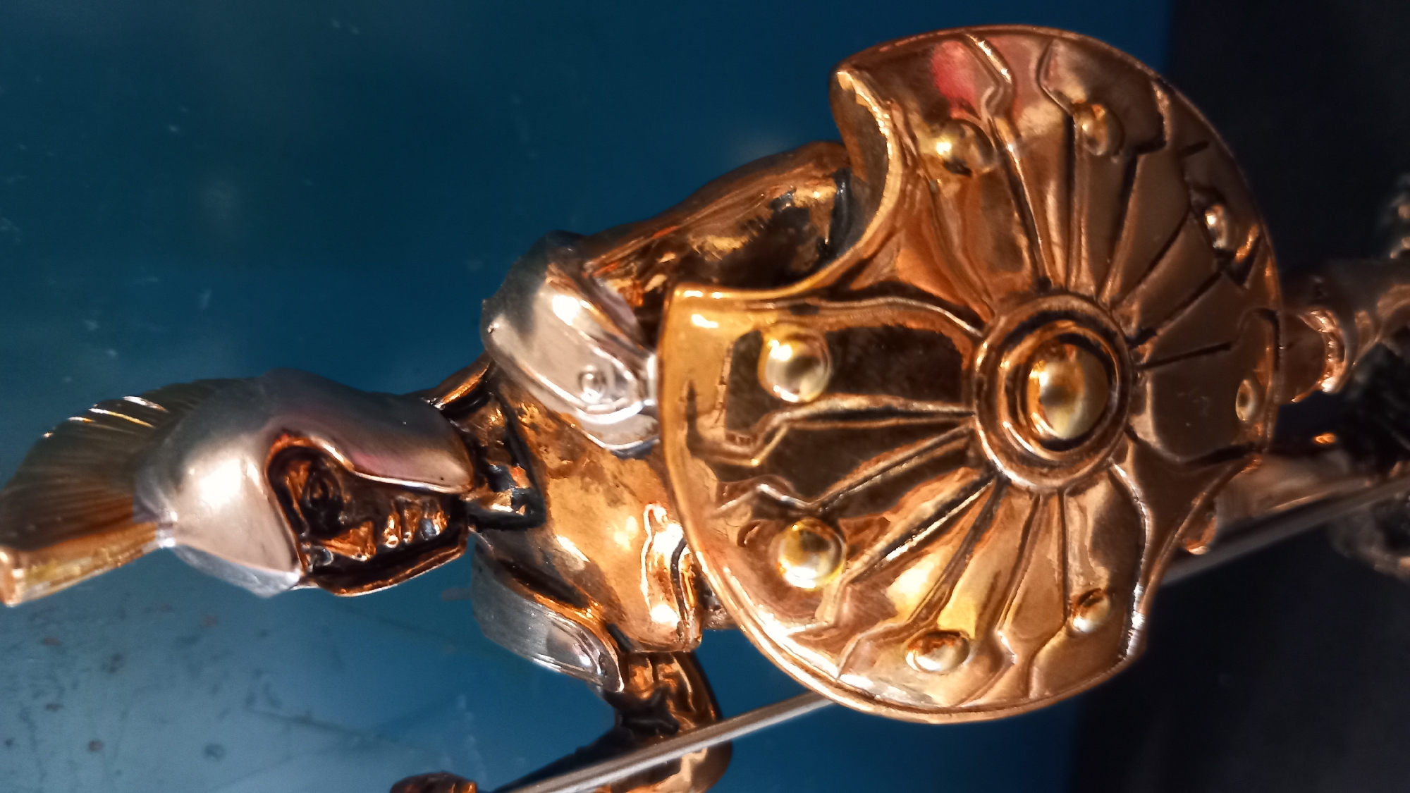 The image shows a close-up of a metallic figurine of a warrior, possibly a Spartan, wearing a helmet with a crest and holding a shield. The figurine is primarily bronze-colored with silver accents on the helmet and shoulder armor. The shield is round and elaborately decorated with raised circular and fan-like patterns. The background is a dark blue surface. There is no indication of the city of Kos, Greece in the image or its immediate context.