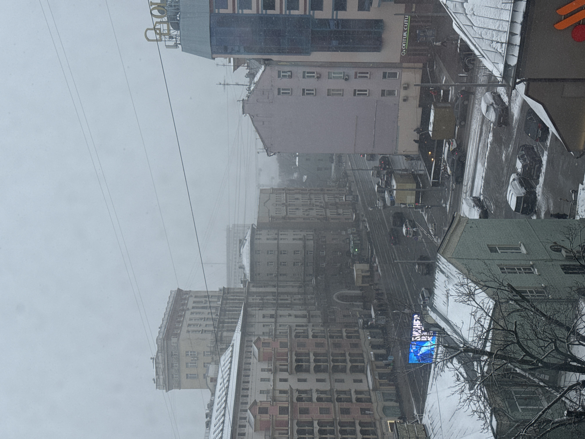This is an urban street scene captured during a snowy winter day in Moscow, Russia. The image is taken from an elevated perspective, looking down and across a city street lined with multi-story buildings.
The dominant feature is the heavy snowfall, with large flakes visible against the overcast, light gray sky and accumulating on surfaces. Snow covers the roofs of buildings and the ground, including the street, which appears slushy or wet from traffic. Visibility is somewhat reduced due to the falling snow.
Numerous buildings of varying architectural styles and heights line both sides of the street. Some buildings appear older, possibly from the Soviet era, with distinct cornices and window arrangements, while others show more modern facades with large glass sections. Air conditioning units are visible on the exterior walls of several buildings. Power lines crisscross the sky, adding to the urban landscape.
The street below is busy with vehicles, primarily cars, driving in both directions. The road surface is dark, likely asphalt, but partially covered in snow and slush. Red traffic lights are visible at an intersection further down the street, indicating active traffic flow.
In the foreground to the right, bare tree branches, devoid of leaves, are partially visible, covered in a light dusting of snow.
Visible text on signs includes:
* On a building to the right, a sign reads "O.OPTİK CUTU".
* Further down the street, on a building, the brand name "ADAMAS" is visible, which is a known jewelry chain in Russia.
* A bright blue LED display on a building in the mid-ground shows fragmented Cyrillic text, including what appears to be "НЬЮ" (New) and other letters suggesting words like "ОДЕЖДА" (Clothing) or "САЛОН" (Salon).
* On a rooftop toward the top of the image, a large golden sign, partially obscured, appears to have Cyrillic letters, possibly spelling out "ТОВАРЫ" (Goods) or a similar word.
The scene conveys the cold, bustling atmosphere of a city enduring a winter snowfall.