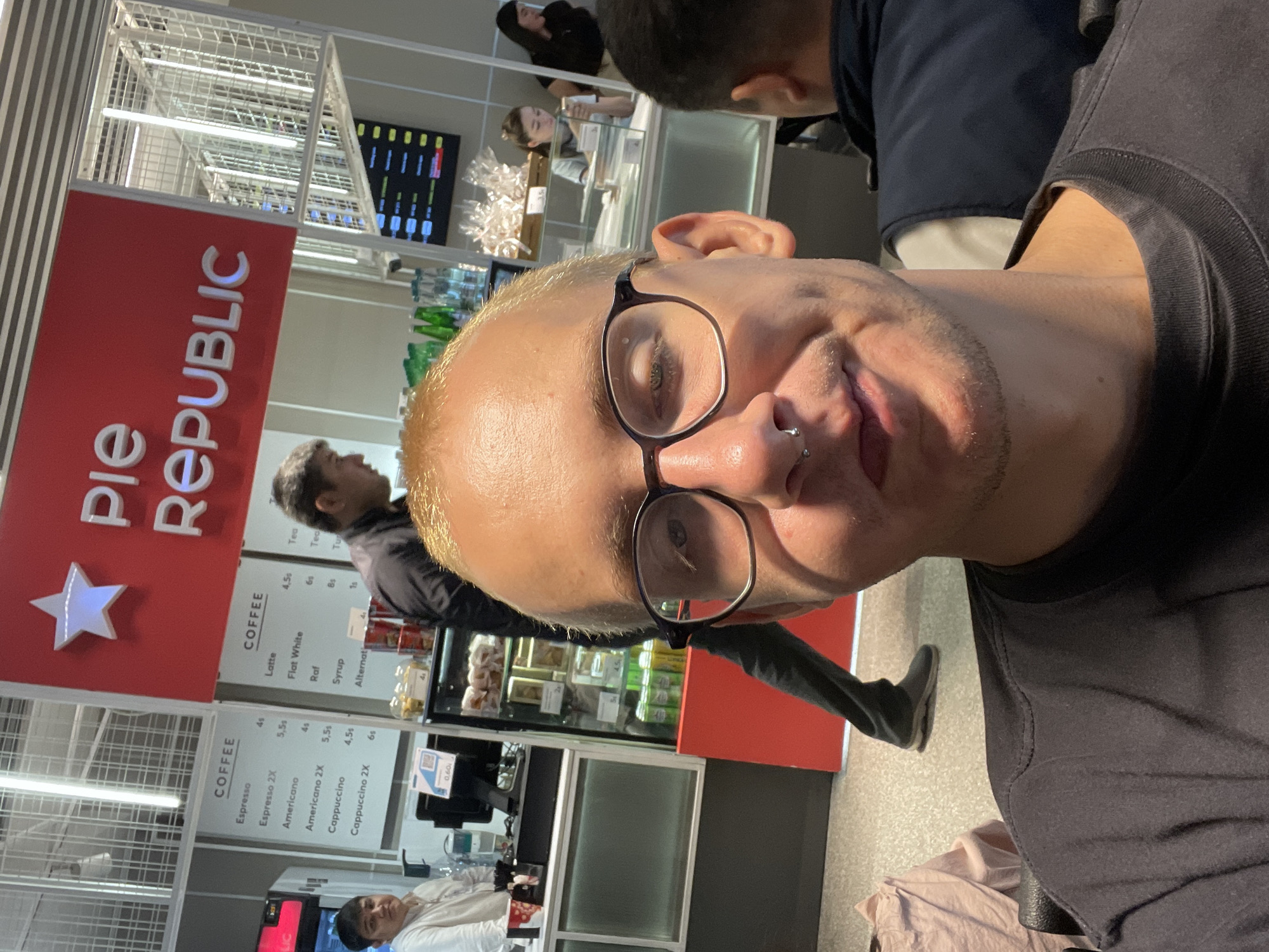 The image is a selfie taken indoors, possibly at an airport or a shopping mall in Tashkent, Uzbekistan. The person taking the selfie is a man with short, blonde hair and glasses. He is wearing a dark grey t-shirt. Behind him is a "Pie Republic" kiosk. The sign is red with white text and a star. The menu to the left lists coffee options like espresso, americano, cappuccino, latte, flat white, and tea with their respective prices in som. There are people standing in line at the kiosk. The floor is tiled and the ceiling has modern lighting fixtures.