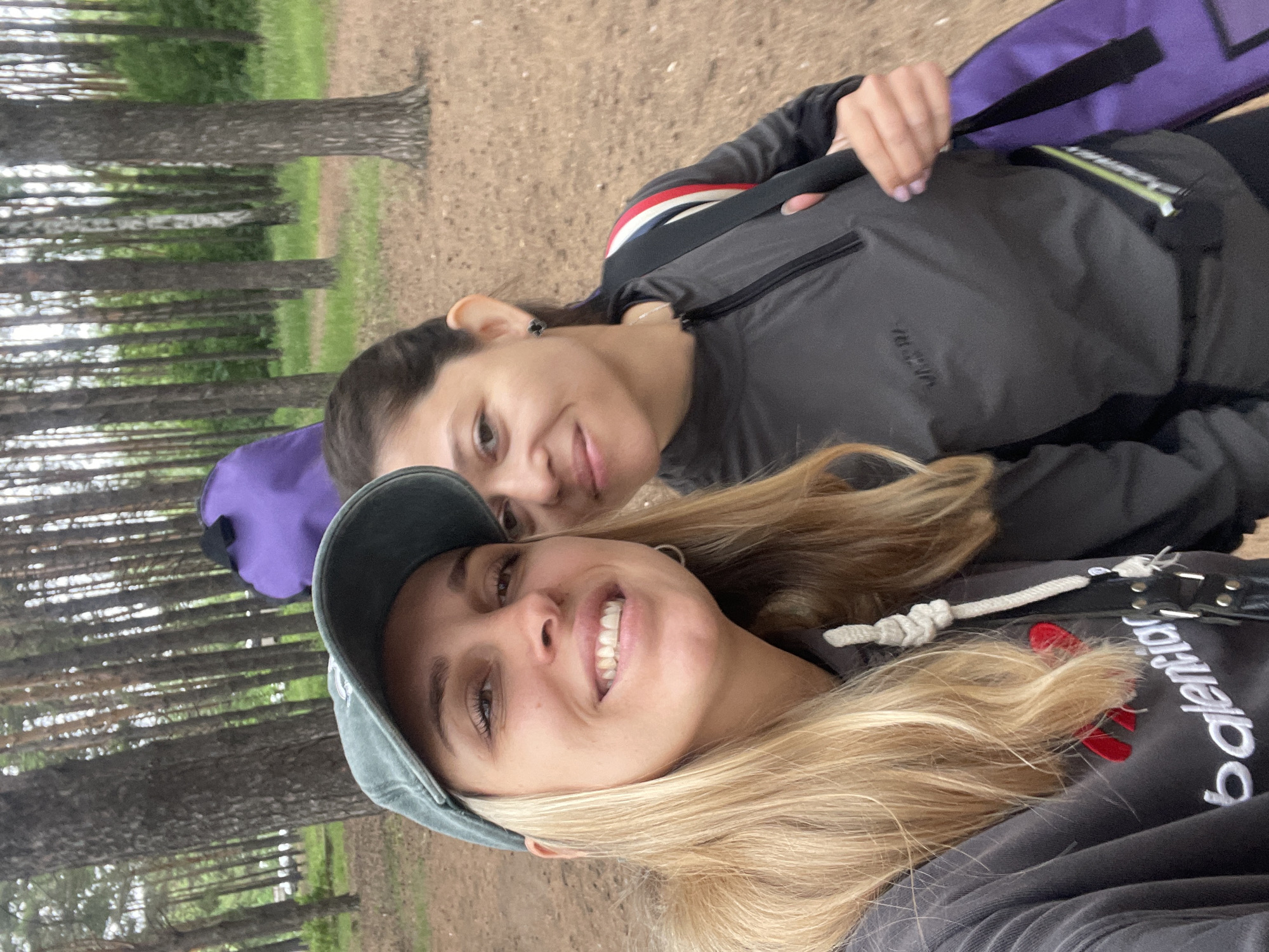 Two women are posing for a selfie outdoors in a forest with tall trees and sandy ground. The woman on the left is wearing a dark hoodie with "balenciaga" printed on it in white, a dark green baseball cap, and has long blonde hair. The woman on the right is wearing a dark grey quarter-zip jacket and has dark hair. Both women are smiling and appear to be enjoying their time. A purple bag is visible behind the woman on the left. The overall impression is one of casual outdoor recreation. The location is identified as Chaikovsky, Russia.