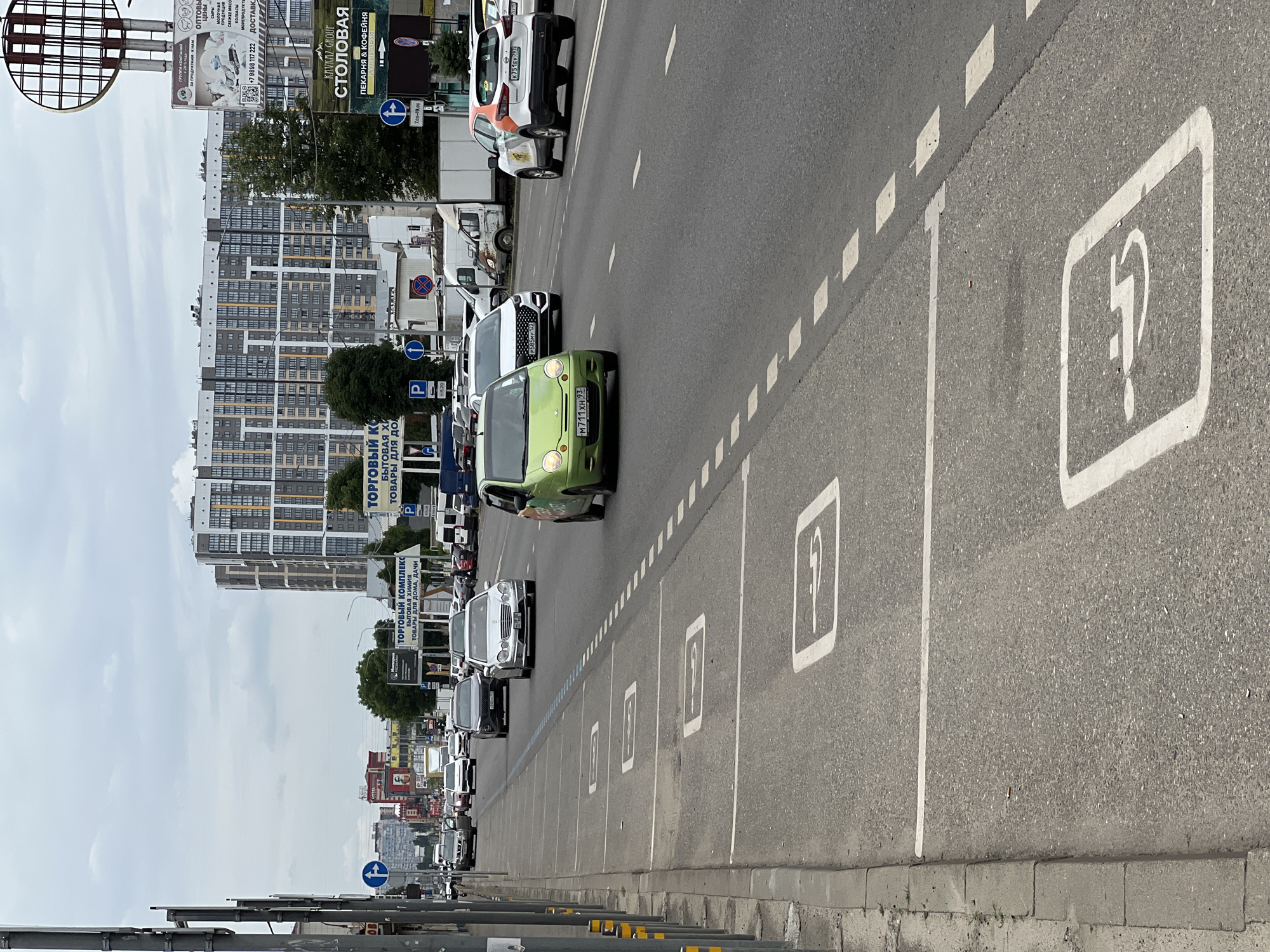The image captures a street scene in Krasnodar, Russia, featuring a multi-lane road with vehicular traffic and designated parking spaces marked with symbols. The overall environment is urban, with tall residential buildings in the background, some commercial signage, and street infrastructure like traffic lights and poles.
Specifically, the foreground shows a curb, a sidewalk, and a stretch of asphalt road. Several rectangular parking bays are marked on the road surface with white lines, each containing a white symbol. These symbols appear to be pictograms, with the most discernible one resembling a person in a wheelchair, indicating accessible parking spots. A few cars are visible on the road, some in motion and others potentially parked or in traffic. One car, a small green hatchback, is in the left lane. Further down the road, more cars are seen, including a silver sedan and a white and orange car.
In the middle ground, the road continues with dashed white lines separating the lanes. More vehicles are present, and various buildings line the street. Commercial signage is prominent, with signs for "ТОРГОВЫЙ КОМПЛЕКС" (Shopping Complex) and "СТОЛОВАЯ" (Canteen) visible. Traffic signals and street lamps are also part of the infrastructure.
The background is dominated by a large, modern residential complex with a distinctive facade featuring multiple windows and contrasting colors. Other buildings, including what appears to be a hotel with a red facade, are also visible. The sky is overcast with clouds. The overall impression is of a busy urban environment on a cloudy day.