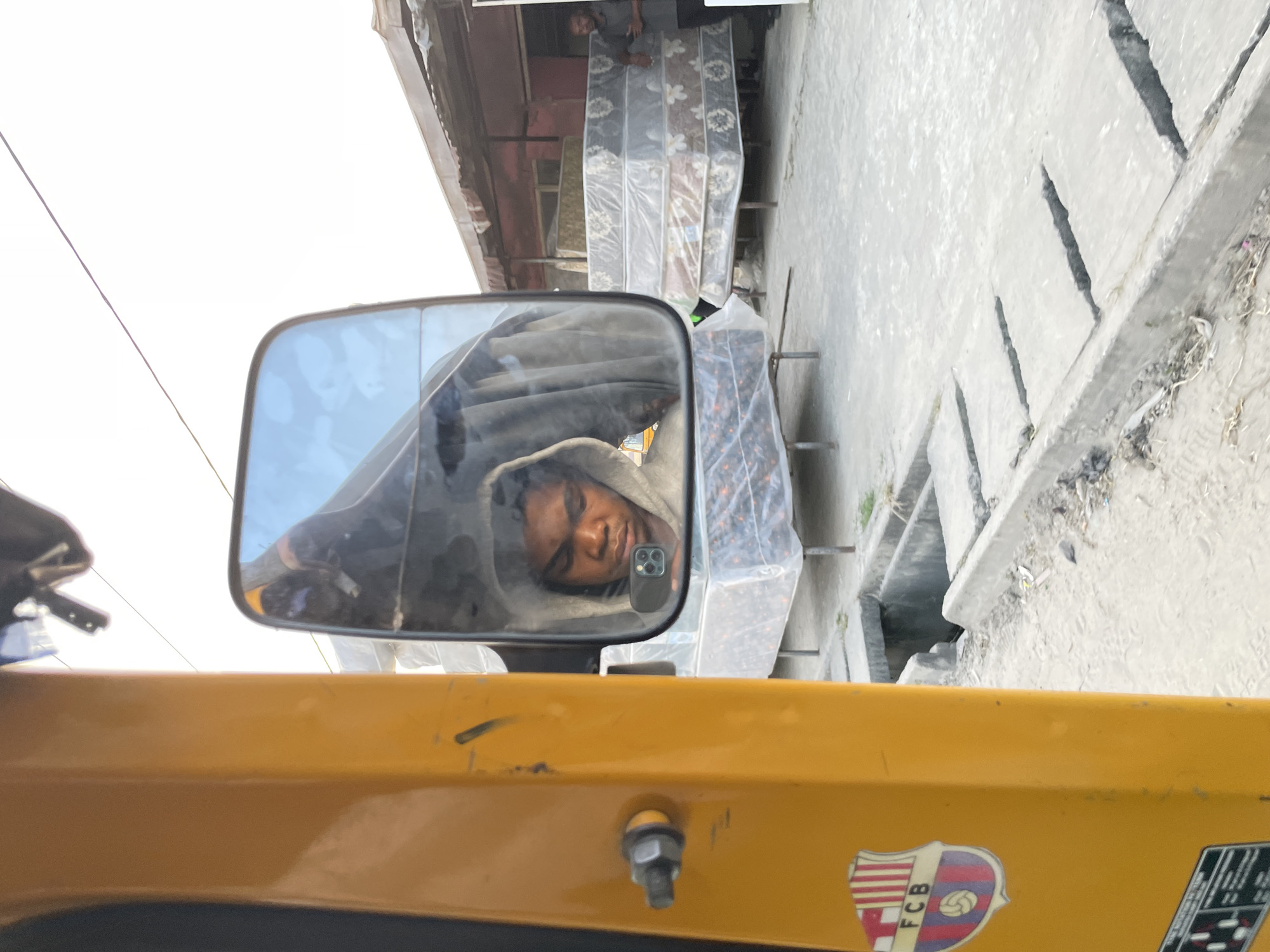 The image, taken in Ado, Nigeria, shows a reflection in the side mirror of a yellow vehicle, likely a tricycle or a similar motorized transport. The reflection captures a young person wearing a grey hooded sweatshirt, holding a smartphone. In the background, through the mirror, several mattresses wrapped in plastic are stacked in what appears to be an outdoor market or a shop. A man in a dark shirt is visible standing next to the mattresses on the right side of the frame. The foreground of the image shows the side of the yellow vehicle with a sticker that resembles the FCB (Futbol Club Barcelona) logo. Also visible are some mechanical parts and a drain grate on the ground. The overall scene suggests a commercial area in the city.