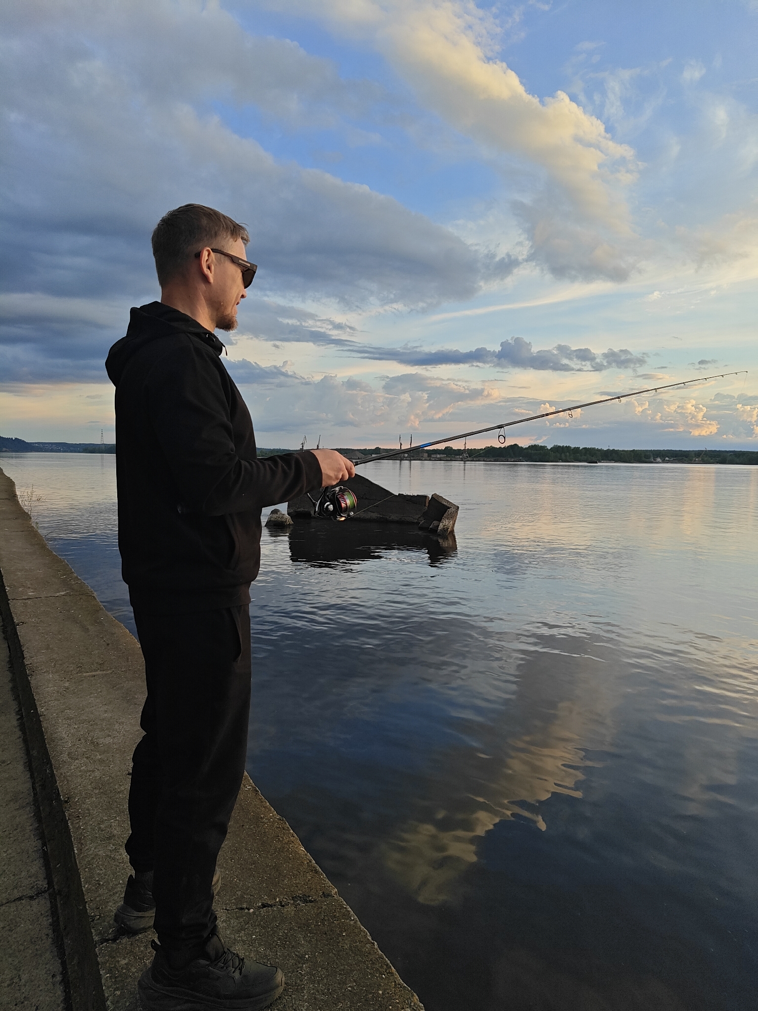 A man wearing sunglasses, a black hooded sweatshirt, black pants, and black shoes stands on a concrete embankment. He is holding a fishing rod and looks out over the water. The water reflects the sky, which is filled with clouds. In the background, there are trees and industrial structures. The location is likely the city of Chaikovsky, Russia.