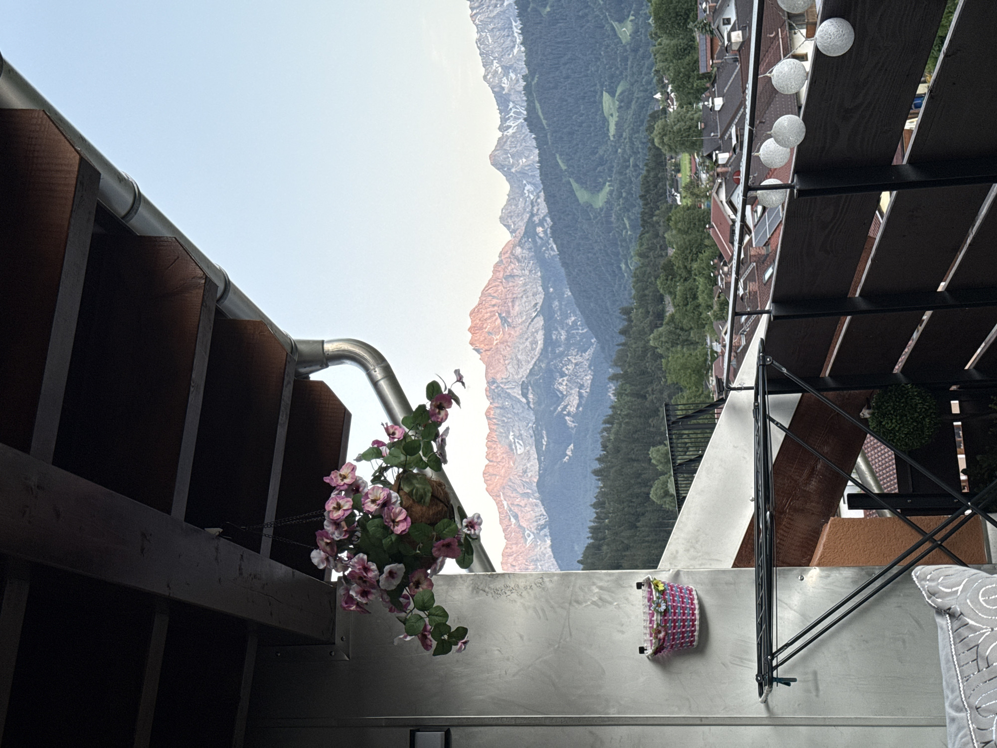 This is a photograph taken from a balcony in Garmisch-Partenkirchen, Germany, overlooking a mountain range and a village. The foreground features a balcony with a wooden railing decorated with string lights. A clothes rack is visible on the left side of the balcony, and a small pink woven basket with flowers is mounted on the wall. Hanging from the balcony roof is a basket of pink and white flowers.
The midground shows a dense green forest that extends to the base of the mountains. Below the forest, a village with red-tiled roofs is nestled amongst the trees. The background is dominated by a majestic mountain range, with the peaks illuminated by the warm glow of the setting or rising sun. The sky is a clear, pale blue. The overall impression is one of a serene and picturesque alpine landscape.