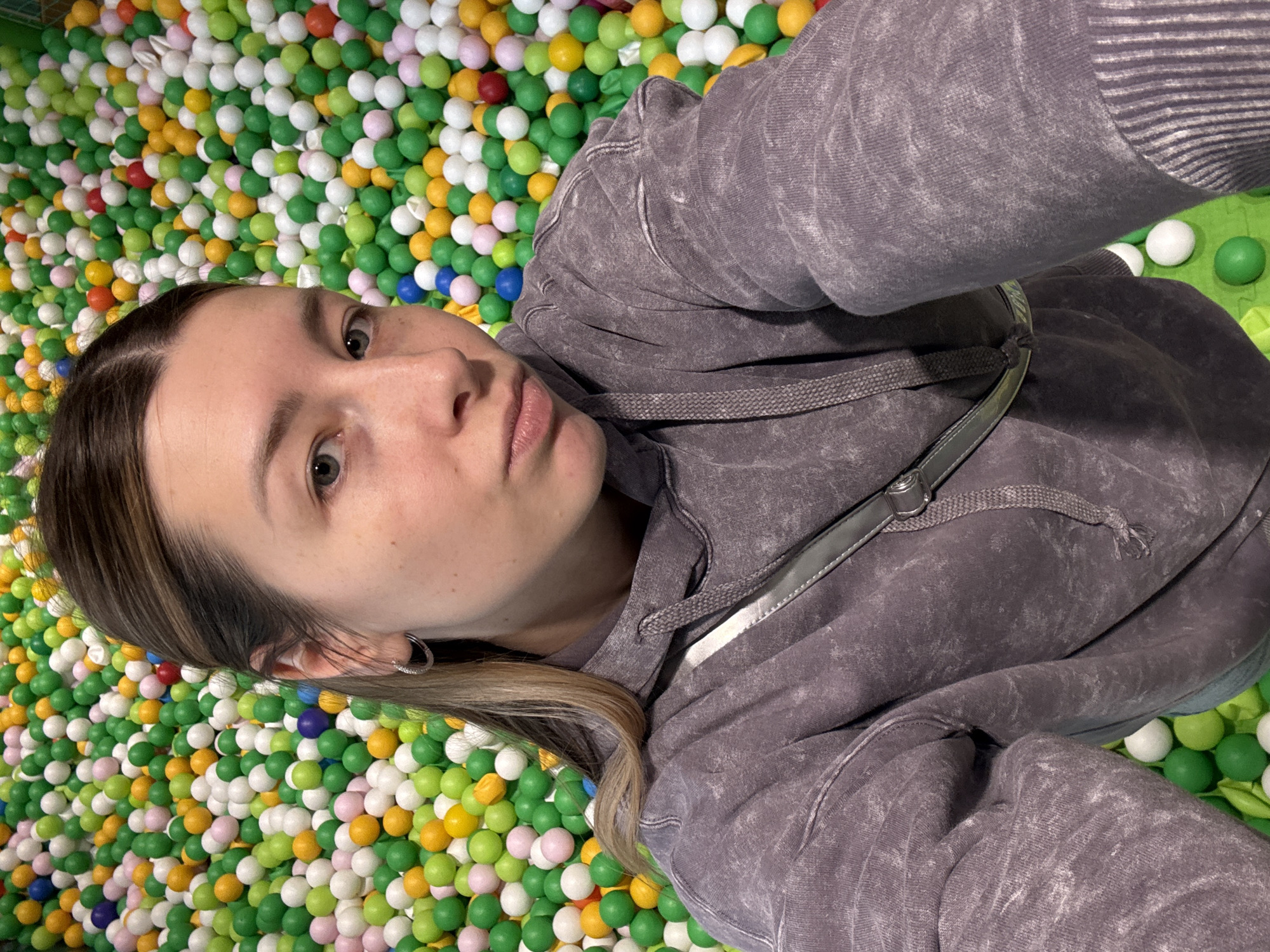 A selfie was taken of a woman lying in a ball pit. The ball pit is filled with green, white, yellow, pink, blue, and red balls. She is wearing a grey acid-washed hoodie with a grey strap over her shoulder. The woman has brown and blonde hair and is wearing a hoop earring. There is green flooring beneath the balls on the right side. The location in Moscow, Russia cannot be determined from the image.