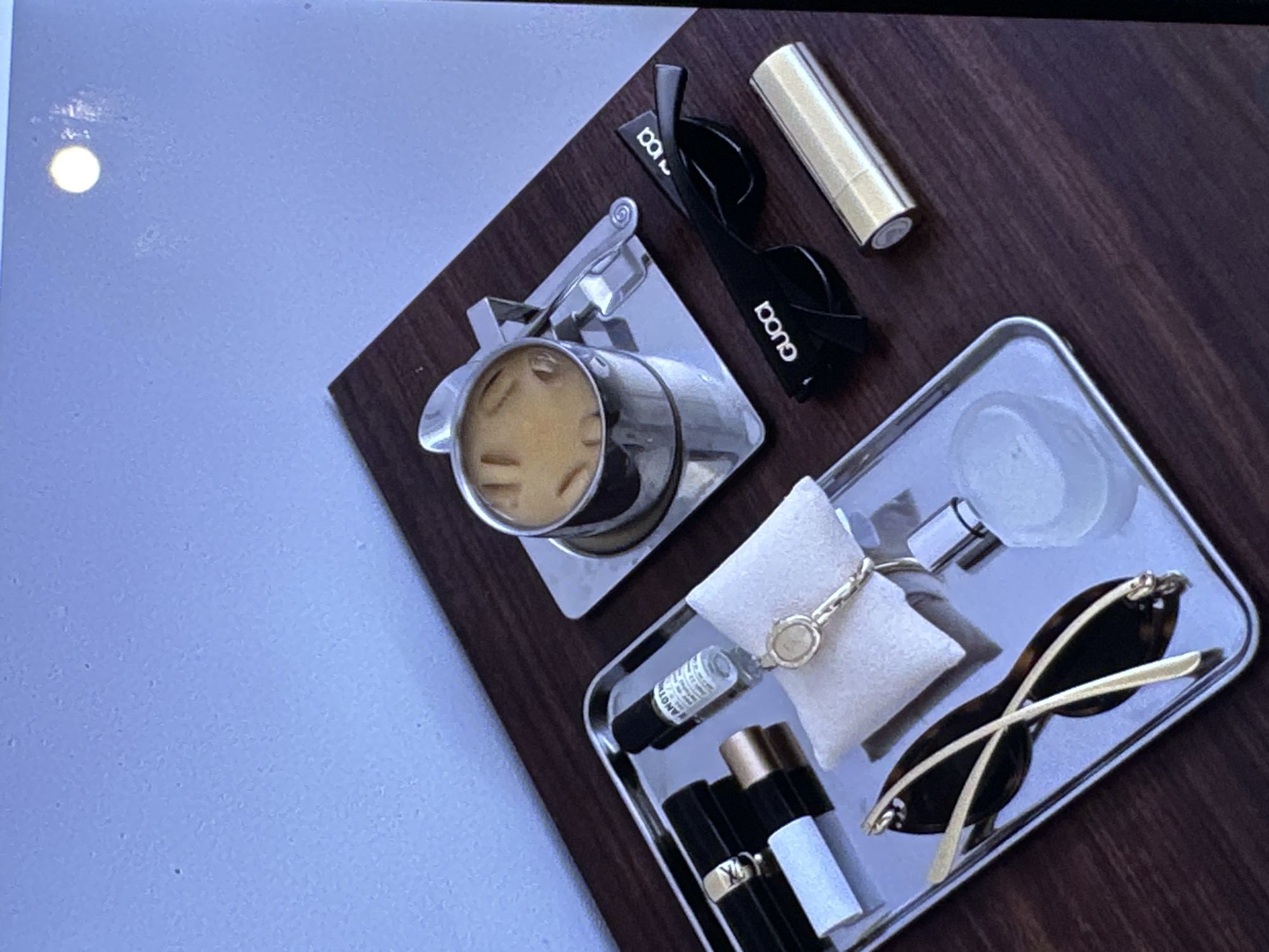 The image presents a flat-lay composition arranged on a dark brown wooden surface. To the left, a stainless steel coffee pot containing a beverage, likely iced coffee, rests on a small, square silver tray. To the right, a larger, rectangular silver tray displays various items. These include: a small white pillow with a gold bracelet draped on it, a small bottle, and a collection of lipsticks in black, gold, and white casings. Additionally, sunglasses are present in both trays: black Gucci sunglasses lie adjacent to the coffee pot, while another pair of brown sunglasses with gold accents are placed on the larger tray. A gold lipstick tube is positioned near the Gucci sunglasses. A clear glass bottle with a silver top rests on the large tray. There is no indication that the photo was taken in Yerevan, Armenia.
