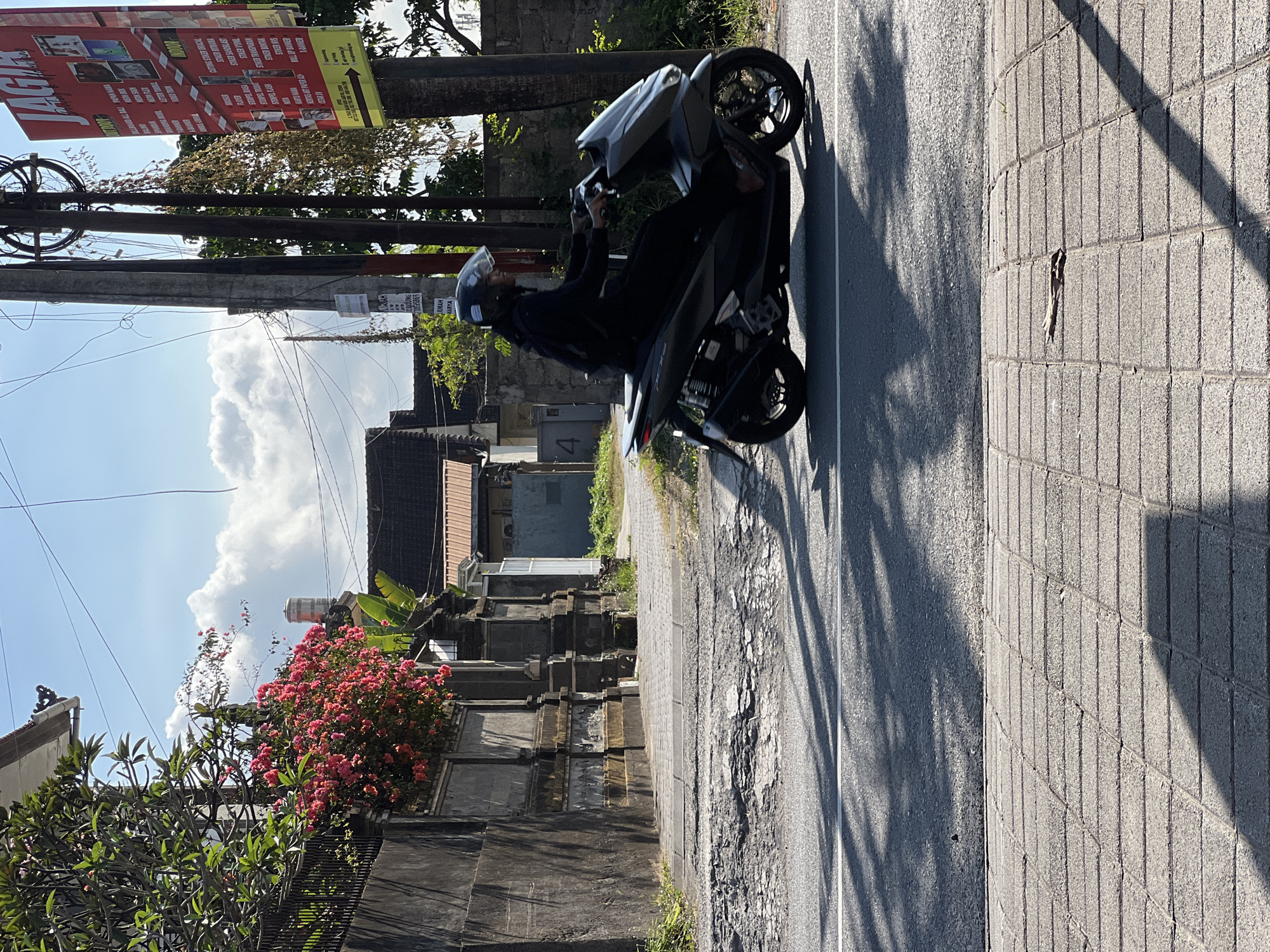 The image captures a street scene in Denpasar, Indonesia, under a bright, partly cloudy sky. A person wearing a black outfit and a white helmet is riding a dark-colored scooter on the right side of a paved road. The scooter is in motion, casting a shadow on the asphalt.
To the left of the road, a weathered concrete wall runs parallel to the street, adorned with decorative stone pillars and topped with lush green foliage and vibrant pink bougainvillea flowers. Beyond the wall, traditional Balinese architecture is visible, with red-tiled roofs. A metal fence is integrated into the wall.
The road itself shows signs of wear, with a section of pavement in the foreground appearing cracked and deteriorated. The sidewalk area is paved with rectangular gray bricks, with shadows from overhead foliage creating a dappled effect. A utility pole with numerous wires stands on the right side of the road, and a brightly colored advertisement poster is attached to it. The poster appears to be in Indonesian, promoting various printing services. The overall impression is a typical, bustling street scene in a tropical urban environment.