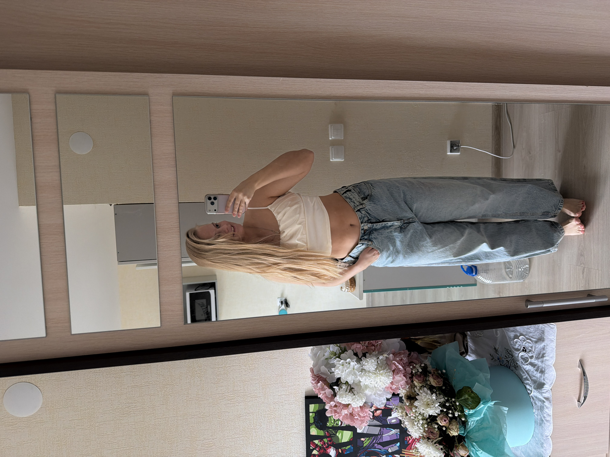 A woman with long blonde hair is taking a mirror selfie indoors. She is wearing a light-colored crop top and light blue jeans. The mirror is part of a closet or wardrobe with multiple mirror panels. The room appears to be a domestic interior, possibly a bedroom, with light-colored walls and flooring. A bouquet of flowers and a framed illustration (possibly anime or comic-related) are visible on a surface in front of the closet. A water bottle is on the floor. There is no indication of location related to the city of Izhevsk, Russia, in the provided image.