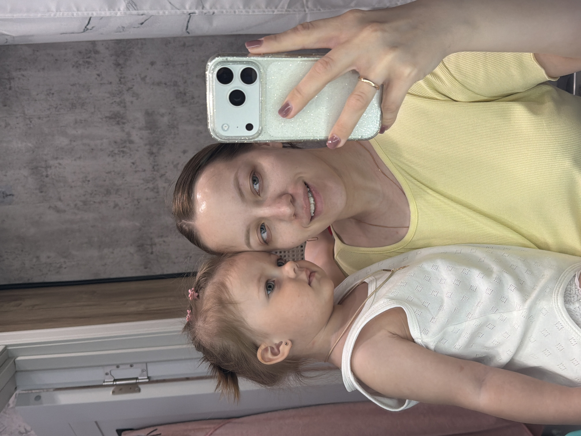 A woman and a young child are taking a selfie indoors. The woman holds a smartphone with a glittery case. The woman is wearing a yellow ribbed top, and the child is wearing a white, sleeveless top with a textured pattern and a thin gold necklace. The background includes a gray textured wall and a window frame. The location is not specified in the image. Therefore, it is impossible to establish that the photo was taken in Reshetnikovo, Russia.