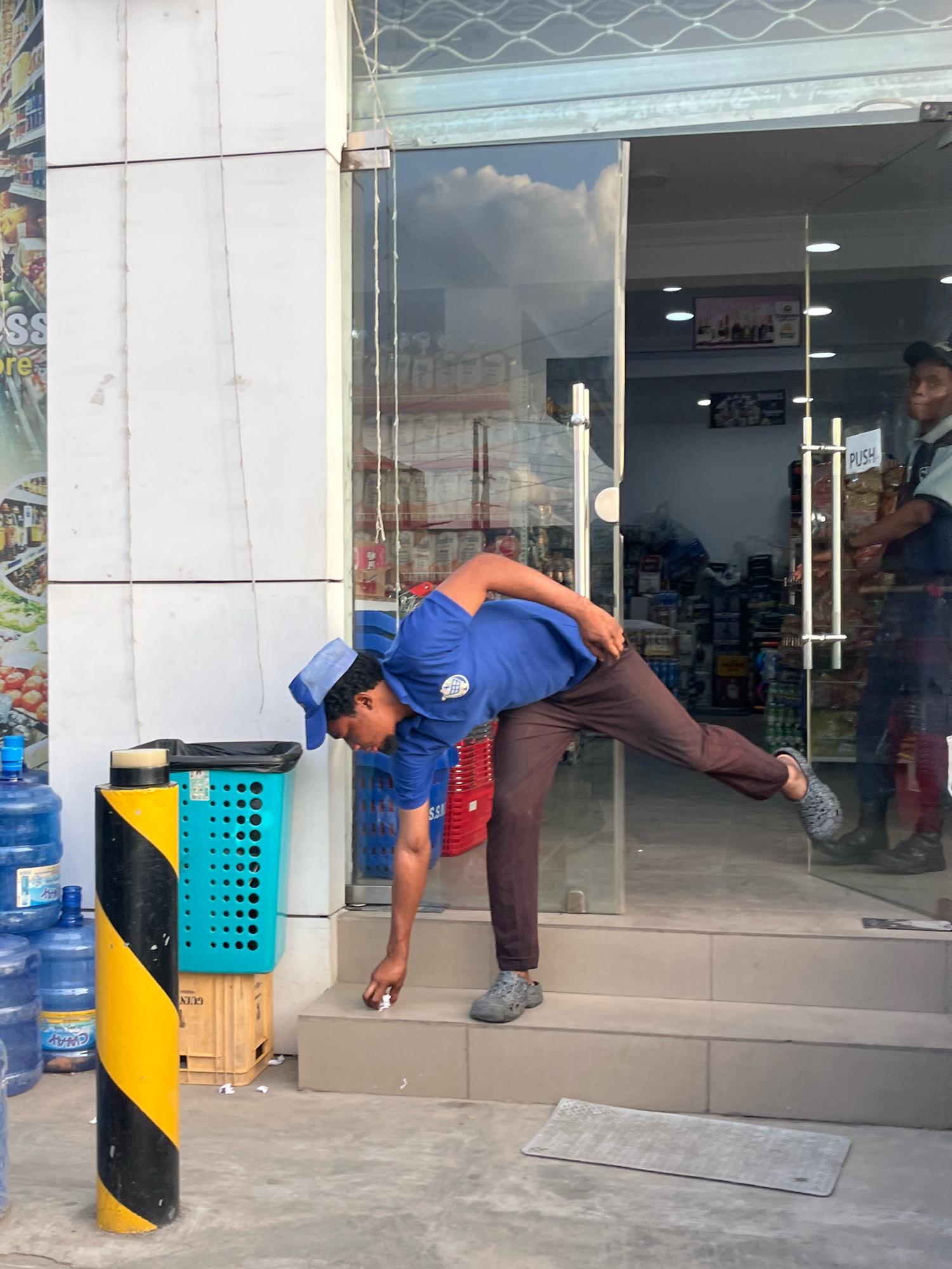 In Ado, Nigeria, an employee wearing a blue shirt and cap is cleaning the entrance to a store. The store appears to be a convenience store or supermarket, with shelves of products visible inside through the glass doors. The employee is bent over, picking up small pieces of paper from the ground with their right hand. They are wearing dark brown pants and grey Croc-like shoes. A blue plastic laundry basket and several large water jugs are stacked to the left of the entrance. A black and yellow striped bollard stands in the foreground. Another individual, possibly a security guard or another employee, is visible inside the store near the entrance, wearing a dark uniform. The ground in front of the store is paved, and there are several steps leading up to the entrance. The overall environment suggests a commercial area in a Nigerian city.