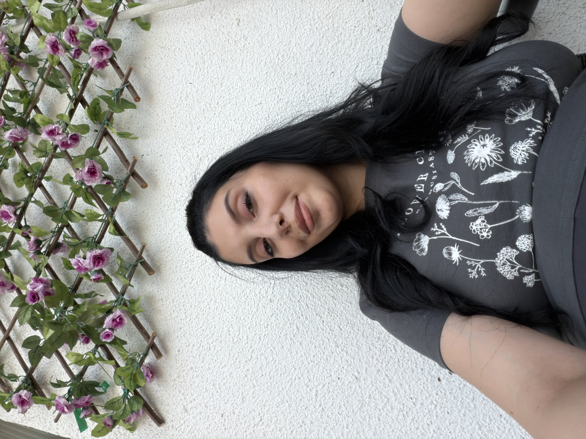 A woman with long, dark hair and light makeup is captured in a close-up selfie. She is wearing a grey t-shirt with a white floral print. She is positioned in front of a white, textured wall, with a wooden lattice decorated with artificial pink flowers and green leaves visible in the upper portion of the frame. The overall setting suggests an outdoor environment, possibly a balcony or garden area. The location is the city of Tbilisi, Georgia.