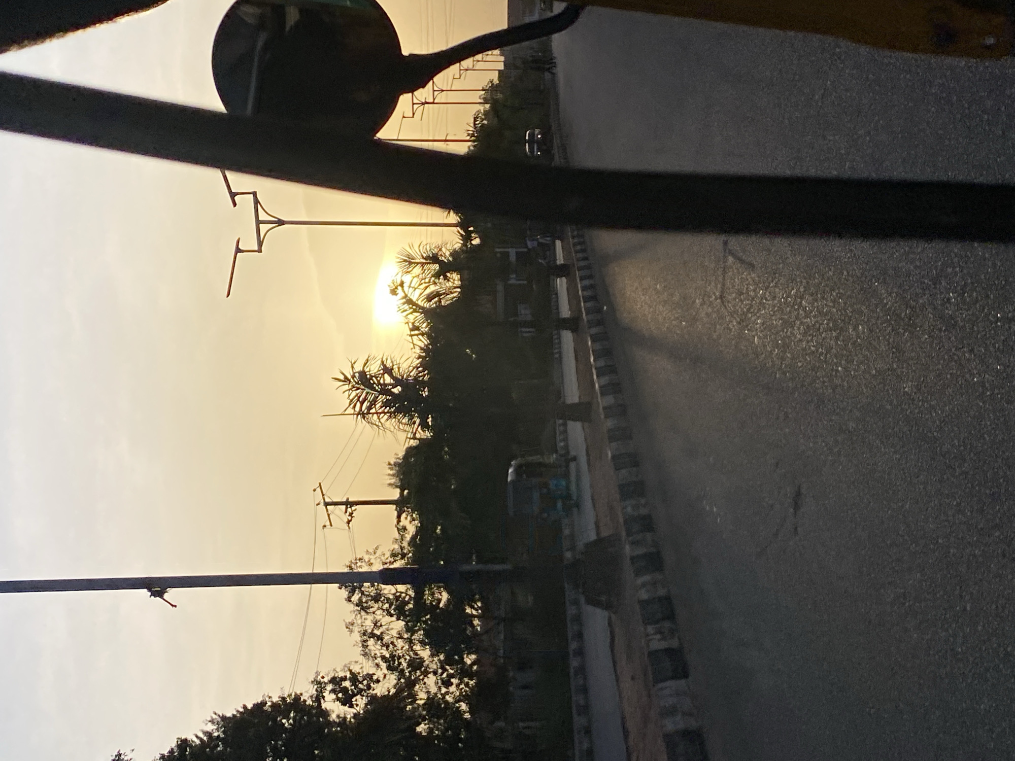 The image, likely taken from a moving vehicle in Barakin Lerawi, Nigeria, shows a road at what appears to be sunset or sunrise. Utility poles line the road. Trees and other vegetation are visible along the roadside. A vehicle, possibly a car, is seen in the distance. The scene is partially obstructed by a side mirror and parts of the vehicle from which the image was captured.