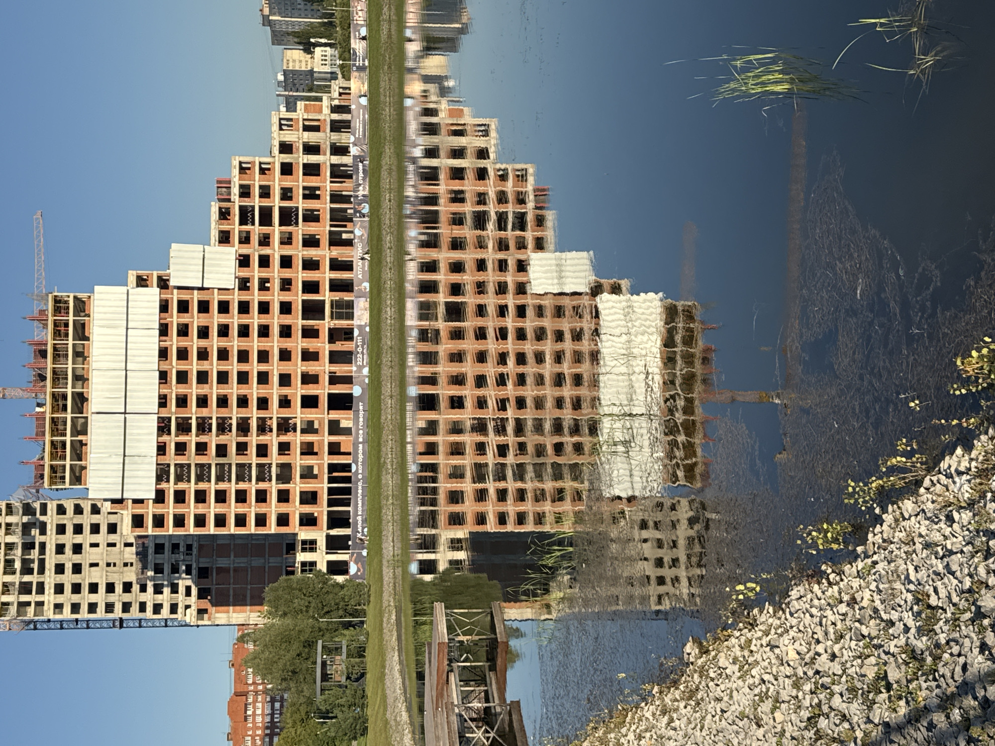 The image captures a construction site in Ufa, Russia, featuring two unfinished buildings reflected in a body of water. The primary building, a multi-story structure with a stepped design, is clad in red brick and concrete, with visible scaffolding and white sheeting. To its left, another taller, unfinished building with a grey concrete facade stands. The reflection in the water distorts the buildings, creating a mirror image with softened edges. In the foreground, a rocky embankment lines the water's edge, with some vegetation growing at the waterline. A wooden dock extends into the water on the left side of the image. The sky is clear and blue, suggesting a sunny day. The overall scene depicts urban development and construction.