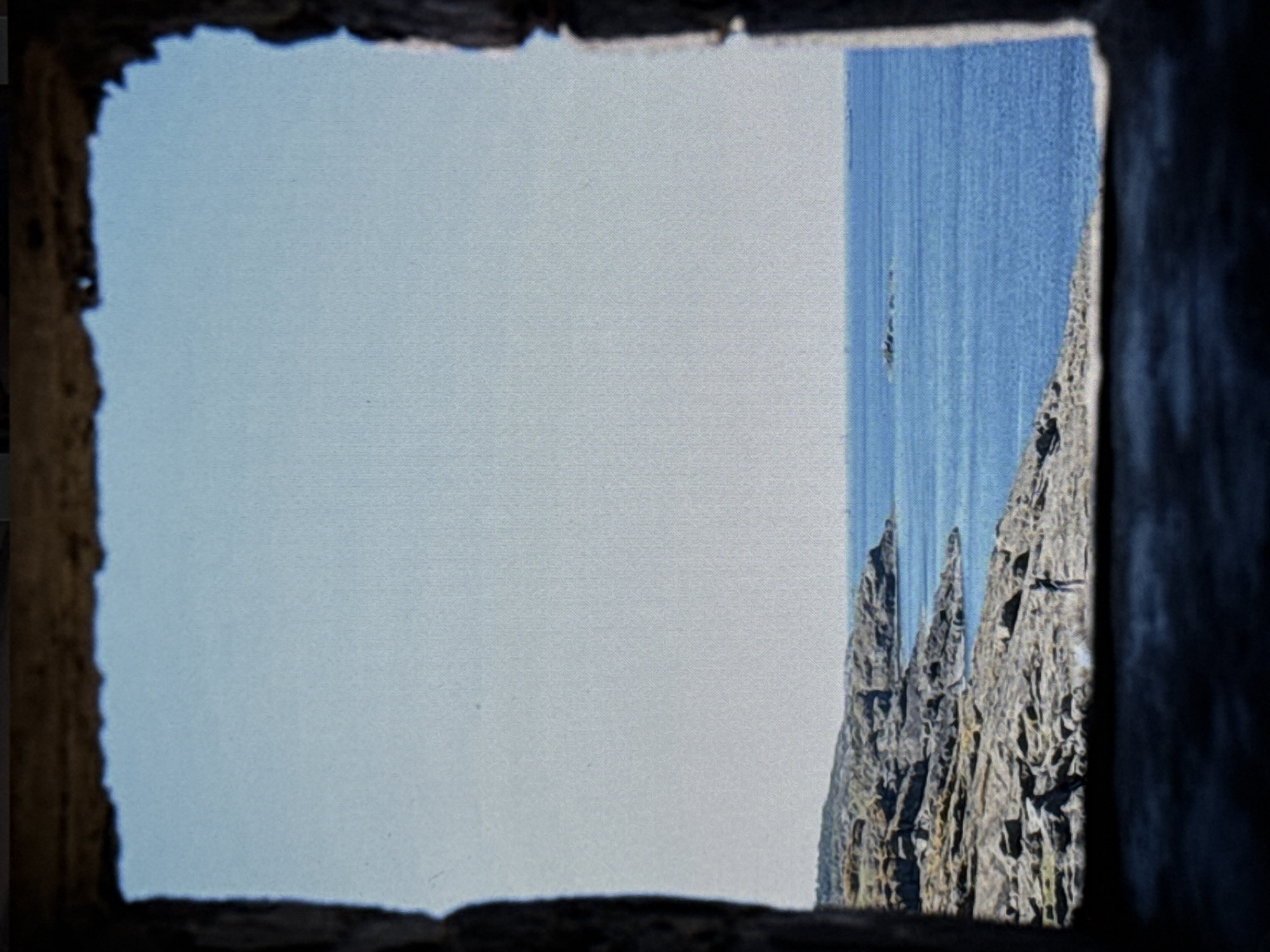 This media file shows a view through a rough, dark opening, possibly a window or doorway, onto a stark landscape. The opening frames a bright, pale blue sky that occupies the left two-thirds of the image. To the right, a section of the deep blue sea is visible, with subtle horizontal lines suggesting waves or currents. Adjacent to the sea, on the right side of the frame, are jagged, rocky cliffs or formations. These rock structures are rough and textured, with dark crevices and lighter, weathered surfaces, giving them a dramatic and imposing appearance. A small, dark figure, possibly a person, is visible on the rocky outcrop. The overall impression is one of a rugged coastal or island environment, viewed from a sheltered, perhaps abandoned, structure. The lighting suggests daytime, with clear skies. The rough framing and the textures of the landscape contribute to a sense of natural grandeur and perhaps isolation. There is no visible text in the image.