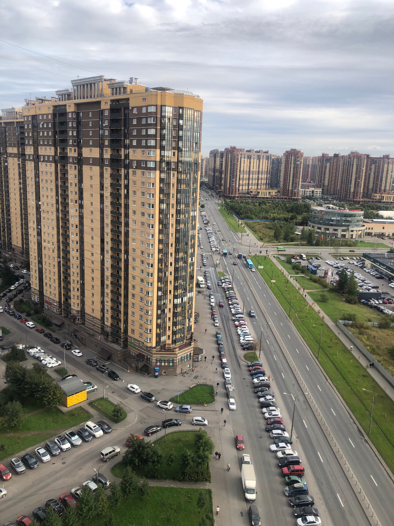 An elevated, wide-angle view captures a dense urban landscape in an unspecified city in Russia. Dominating the foreground is a tall, modern residential building with a beige facade and dark vertical accents, featuring numerous windows and balconies. Adjacent to this building is a multi-lane road filled with parked and moving vehicles, including cars, vans, and a bus. The road stretches into the distance, lined with trees and grassy areas. Across the road, a cluster of similar high-rise apartment buildings forms the middle ground, their architectural styles echoing the foreground structure. Further into the distance, more residential buildings and some commercial structures are visible under a cloudy sky. Several individuals are depicted walking on sidewalks and crossing streets. The overall impression is one of a developed, bustling urban environment.