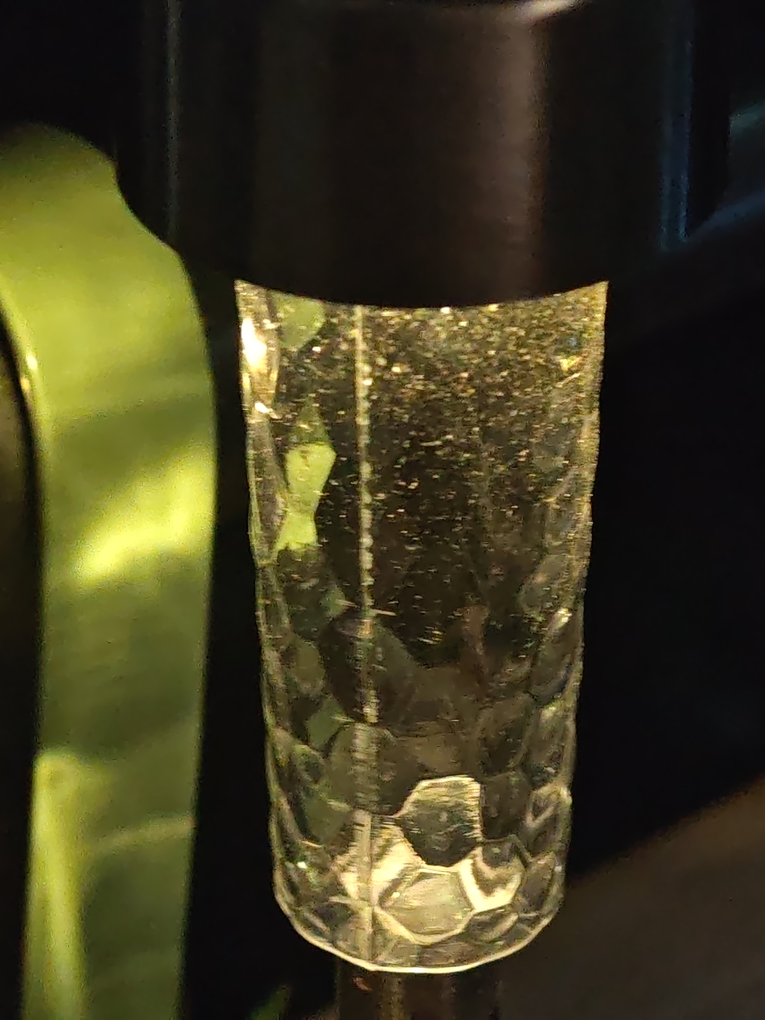 The image displays a close-up, vertical orientation of a translucent, clear object with a textured, faceted surface, resembling hammered glass or plastic. This cylindrical object contains numerous small, bright, scattered reflections or particulate matter, illuminated from within or behind, creating a shimmering effect. It is capped at the top by a dark, opaque, cylindrical or rectangular fixture. To the left, a bright, curved, smooth, olive-green object or surface is partially visible, reflecting light and contrasting sharply with the darker background. The overall background is dark and indistinct, suggesting low ambient light or selective focus. The image contains no visual information, such as landmarks, text, or environmental context, to indicate a location within the city of Shumen, Bulgaria.