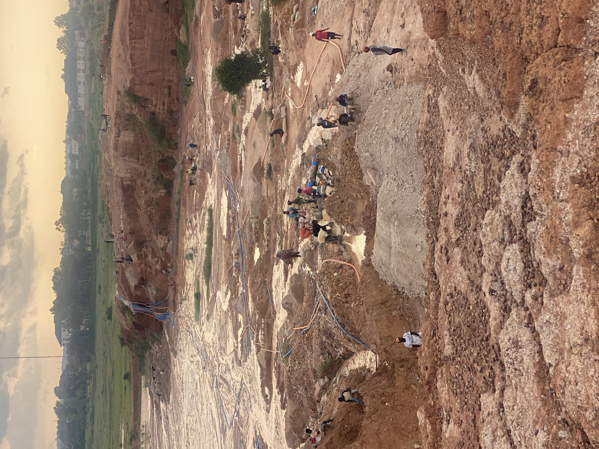 The image depicts an expansive, open-pit mining operation in Jos, Nigeria. The landscape is characterized by a network of muddy channels and excavated earth, indicative of extensive resource extraction. Numerous individuals, appearing to be laborers, are actively engaged in mining activities throughout the scene. They are spread across various levels of the pit, some digging in the soil, others carrying materials, and some gathered near water sources. The environment is marked by reddish-brown soil, piles of excavated dirt, and several orange and blue pipes that seem to be involved in water management or extraction. In the background, the scene transitions to a more vegetated area with scattered buildings and trees, suggesting the mine is located on the outskirts of a more developed area. The sky above is overcast with a soft, diffused light, indicating either early morning or late afternoon.