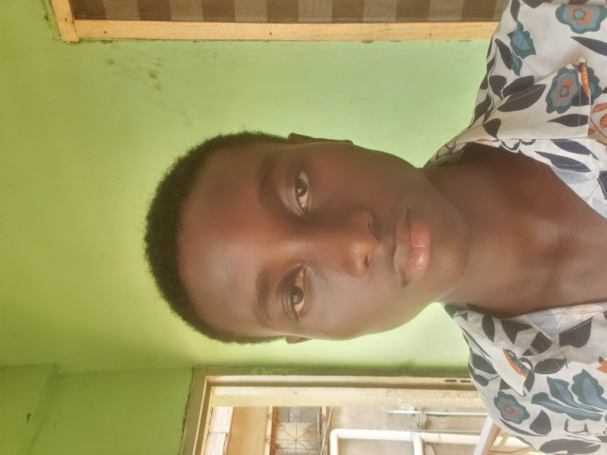A portrait of a dark-skinned person with short, dark hair is in the foreground. The person is wearing a light-colored shirt with a floral pattern. The background consists of a green wall and a doorway leading to an outdoor area. The location is suggested to be Alalubosa, Nigeria, based on prior context. The image appears to be taken indoors.