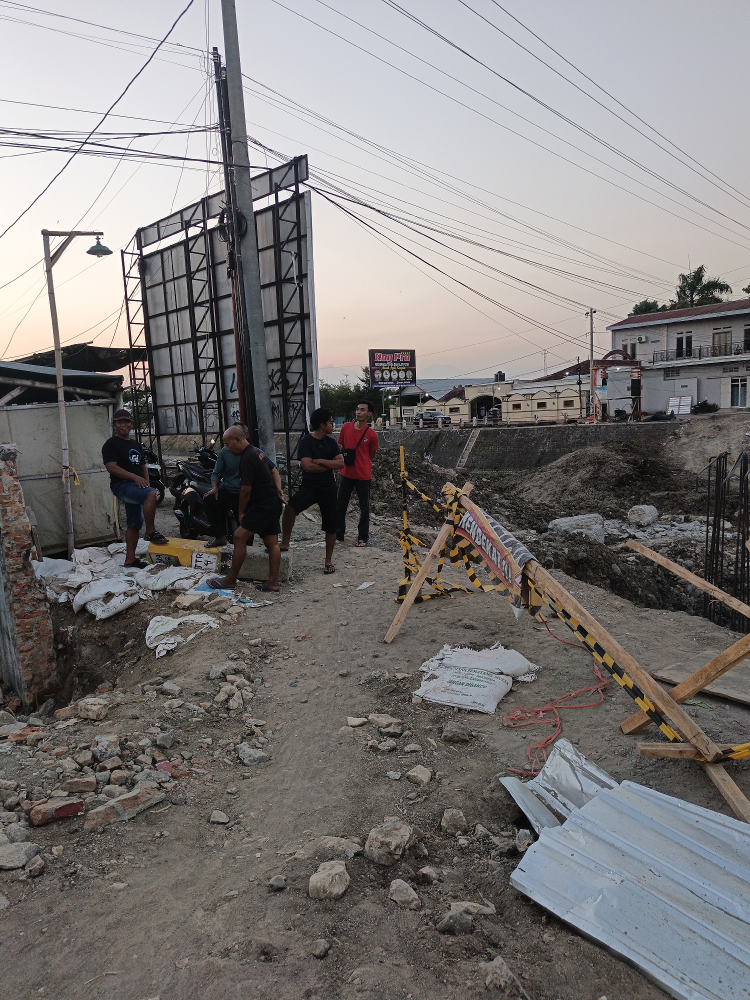 The image depicts a construction site in Planggu, Indonesia, at dusk. Five men are present, seemingly observing the work or taking a break. The foreground is cluttered with construction debris, including bricks, rocks, and discarded materials like white sacks and corrugated metal sheets. A yellow and black striped barrier, labeled with Indonesian text including "AWAS!" and "JANGAN DIGANGGU," indicates a hazardous area. In the midground, a large billboard structure stands behind a utility pole adorned with numerous electrical wires. Beyond the construction site, buildings and trees are visible under a pale, hazy sky. The overall scene suggests ongoing development and infrastructure work.