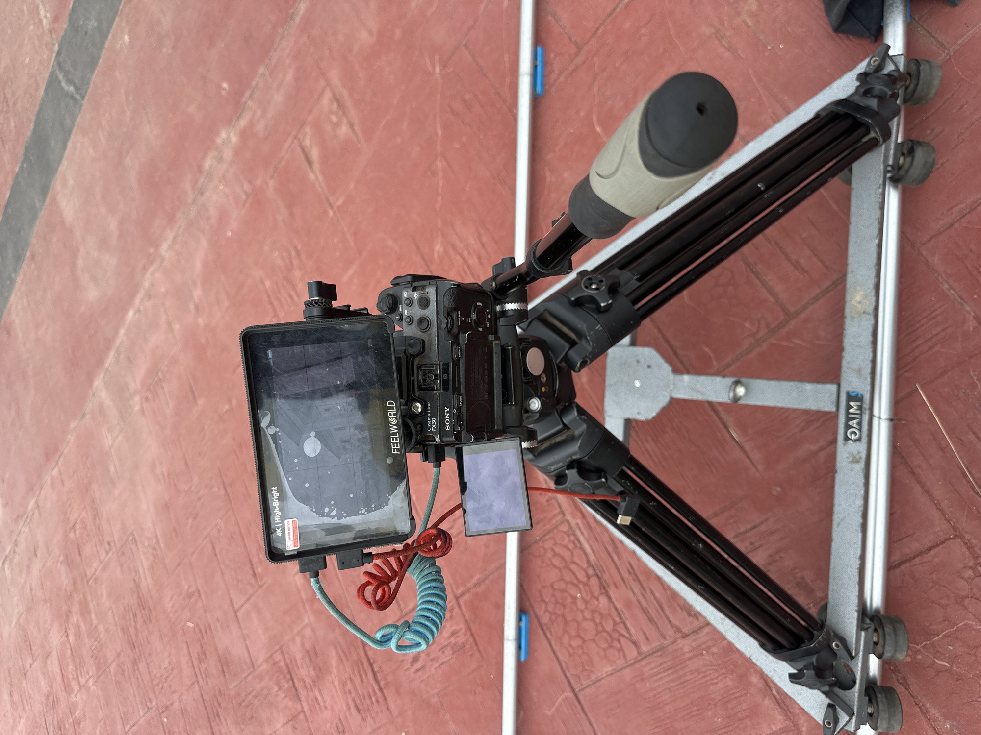 The image shows a Sony FX30 camera with a FEELWORLD monitor mounted on a tripod slider. The camera and monitor are set up on a red, brick-like paved surface. Two coiled cables, one blue and one red, are connected to the monitor. The slider has silver metal rails and black legs, with wheels visible at the bottom. The background is a continuation of the red paved surface. No people are visible in the image. The scene appears to be outdoors, possibly in a public area.