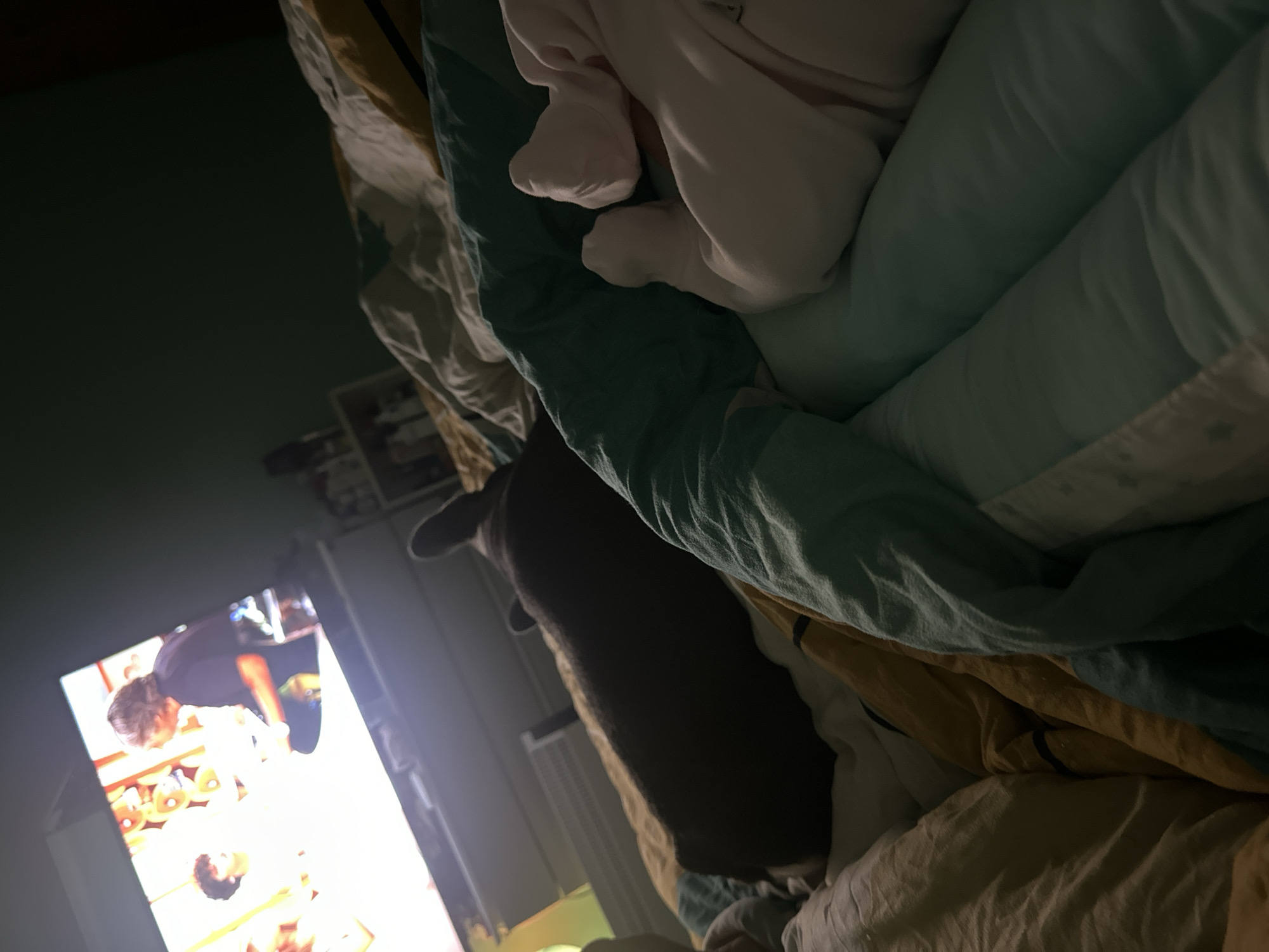 The image captures an indoor scene, likely a bedroom or living room, with dim ambient lighting suggesting nighttime. The perspective is low, looking upwards and across a bed or couch.
In the foreground, a medley of crumpled bedding and blankets in various shades of beige, yellow, light blue, and teal occupies the right and top portions of the frame. Within these covers, a light pink or white item resembles a soft onesie or blanket, with two small, bundled shapes suggesting baby feet or tiny human feet tucked inside. This implies a person, possibly a child or baby, is nestled under the blankets.
To the left of the bedding, a dark brown object, possibly a pet such as a dog or cat, is partially visible, lying down and facing towards the left side of the image.
The left side of the frame is dominated by a brightly illuminated television screen displaying content. Two human figures are visible on the screen: one in a dark shirt standing and leaning over, and another person lying down, viewed from the chest up with dark hair. The specific activity on the screen is unclear due to the bright light. Behind the television, some dark, out-of-focus shelves or furniture can be seen against a dark green wall.
The overall scene conveys a sense of comfort and relaxation, with someone cozy under blankets, potentially with a pet, watching television. There is no visible text.