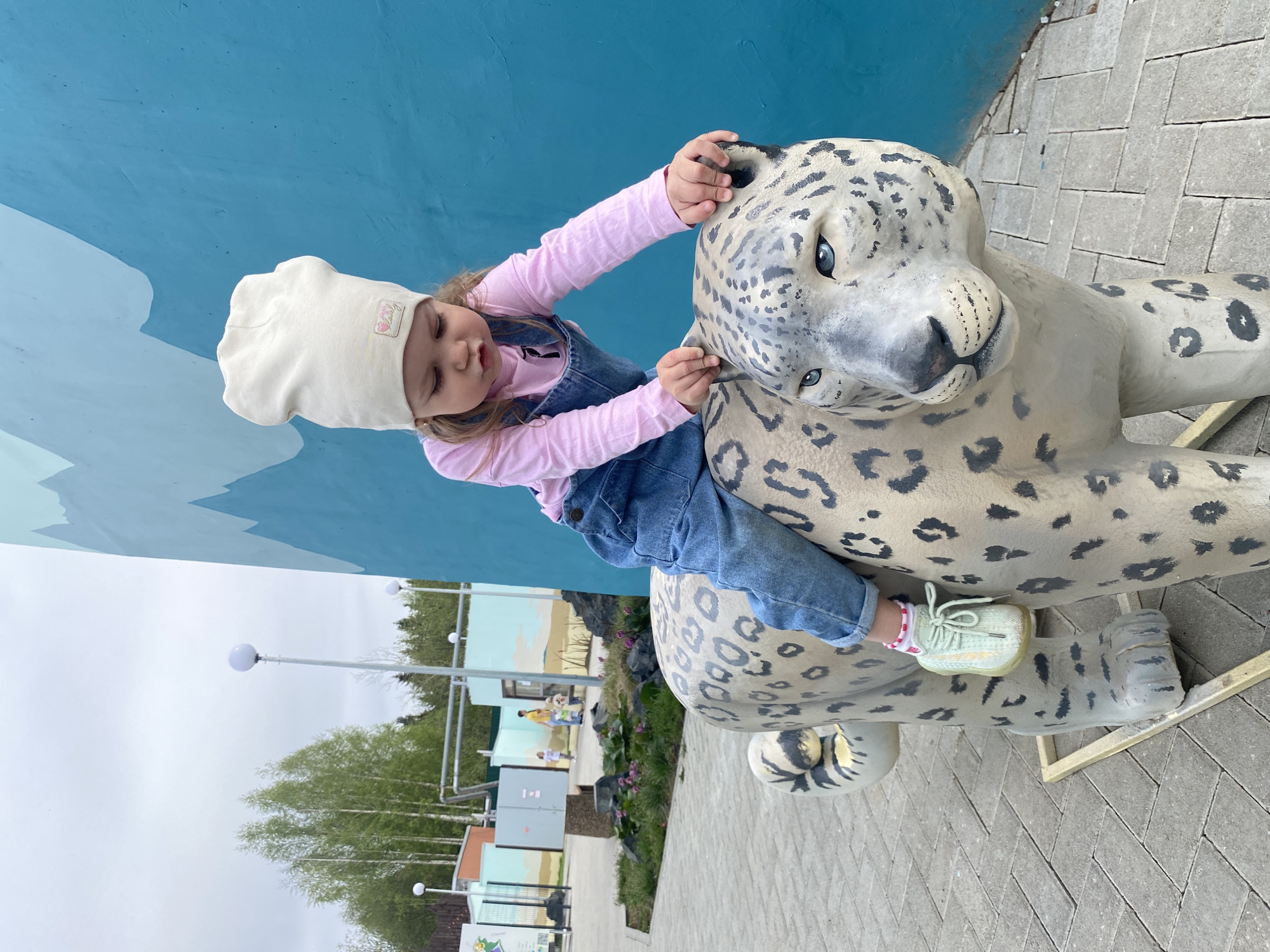 A young girl in a pink long-sleeved shirt, denim overalls, a cream-colored hat, and light green sneakers is sitting on a large, spotted snow leopard statue. The girl has her hands on the leopard's head, appearing to be petting it. The statue is placed on a paved area with a herringbone pattern. Behind the statue and the girl is a blue wall and scenery with green trees, bushes, and structures.
The location is the Perm Zoo in Perm, Russia. This is evident from the layout and design, which matches publicly available images of the zoo.