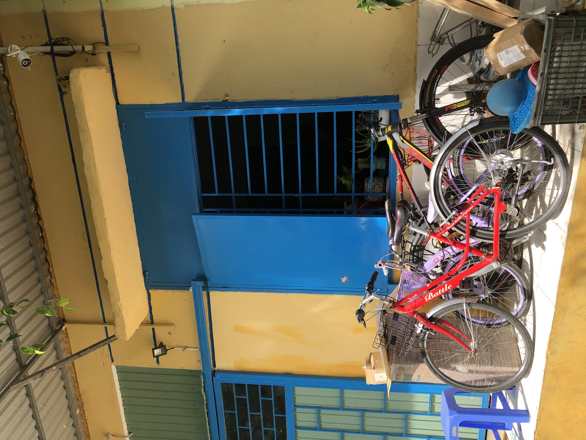 The image shows a collection of bicycles leaning against a yellow and blue building, likely in an outdoor space. The bicycles are of various colors, including red, purple, and black. One red bicycle has a basket in the front and the word "Battle" is visible on its frame. Another black bicycle is partially visible behind the red one. A blue plastic stool is placed in front of the bicycles. There are also some boxes and a blue basket near the bicycles. The building has a blue door with metal bars and a yellow wall. A security camera is mounted on a white pipe near the top left of the image. The lighting suggests it might be daytime, with some shadows indicating the sun's position. The scene appears to be in a residential or commercial area in Da Nang, Vietnam, given the building style and presence of multiple bicycles. There are no people visible in the image.