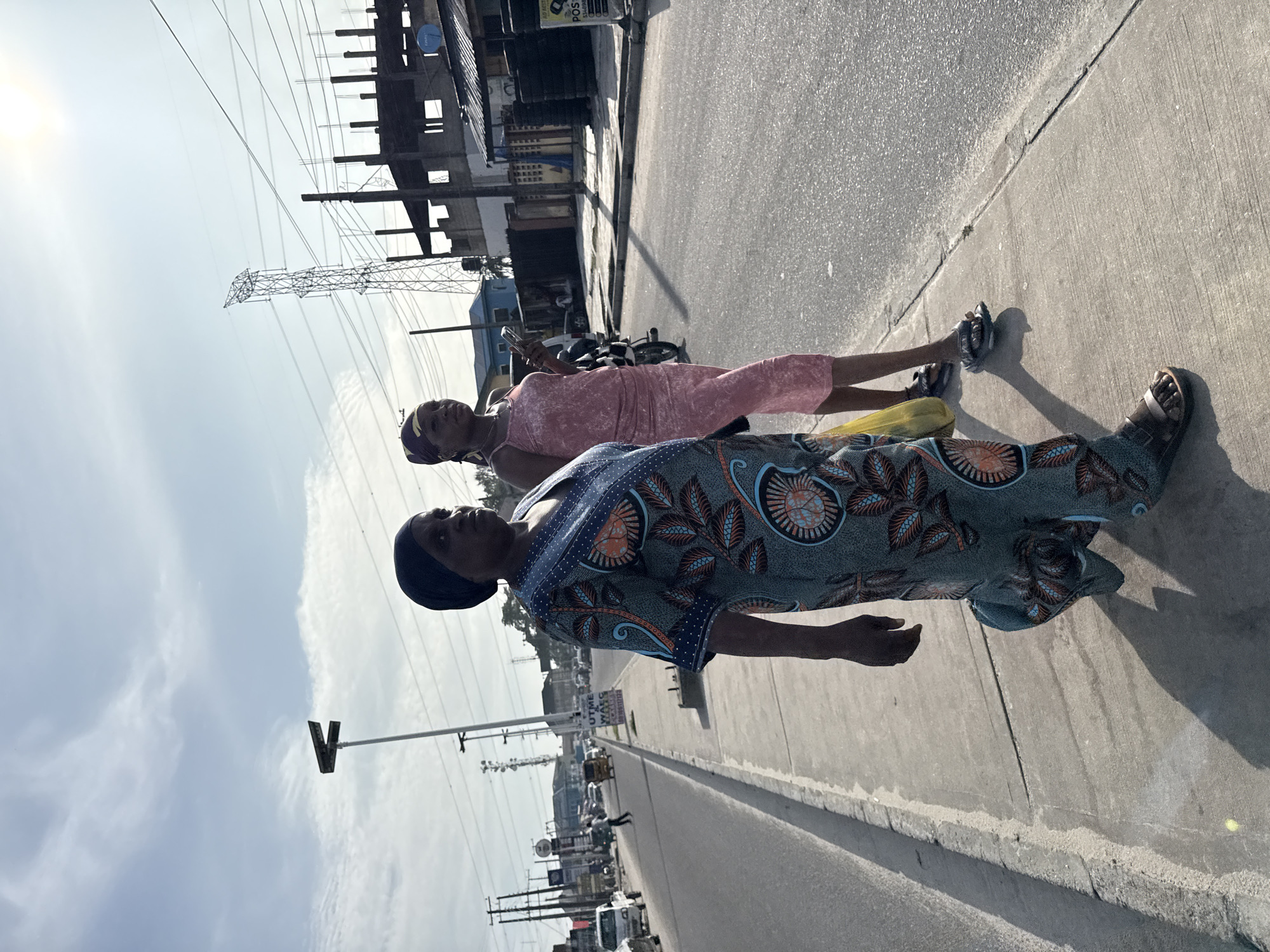 Two women stand on a concrete sidewalk next to a road in Ado, Nigeria. One woman wears a patterned blue and orange dress and the other wears a pink dress. Both women wear head coverings. A motorcycle is parked on the road behind them. In the background are buildings, power lines, and vehicular traffic. The sky is visible with light clouds.