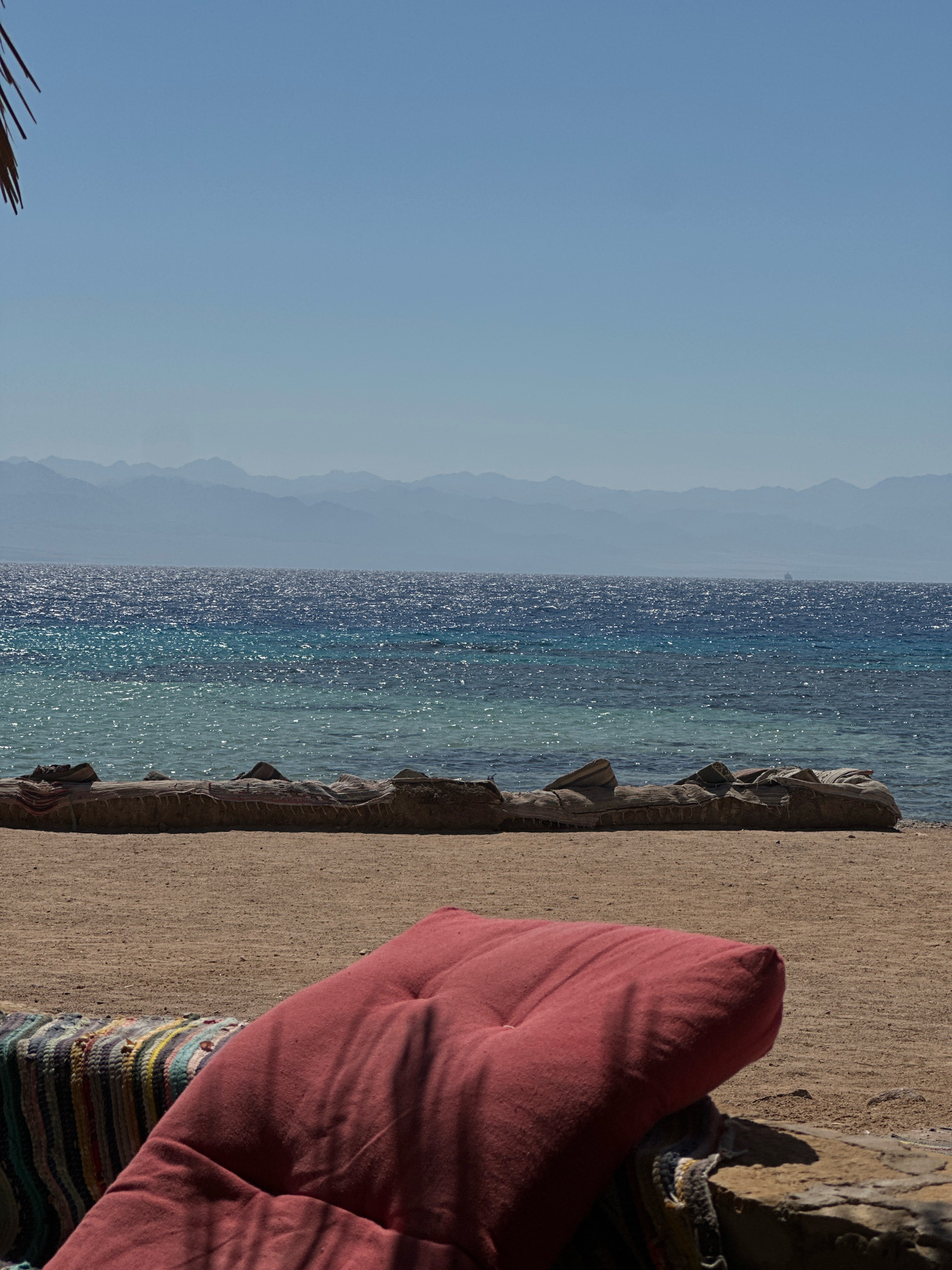 The image depicts a serene beach scene in Nuweiba, Egypt, under a clear blue sky. In the foreground, a large, deep red cushion rests on a stone ledge, partially obscuring a colorful striped woven textile. The beach itself is sandy, with a line of rocks and debris situated near the water's edge. The water is a vibrant turquoise transitioning to a deep blue, shimmering with sunlight, and a distant mountain range, possibly in Saudi Arabia, is visible across the sea, softened by atmospheric haze. A portion of a palm frond is visible in the upper left corner, suggesting a tropical or subtropical environment. The overall impression is one of tranquility and natural beauty.