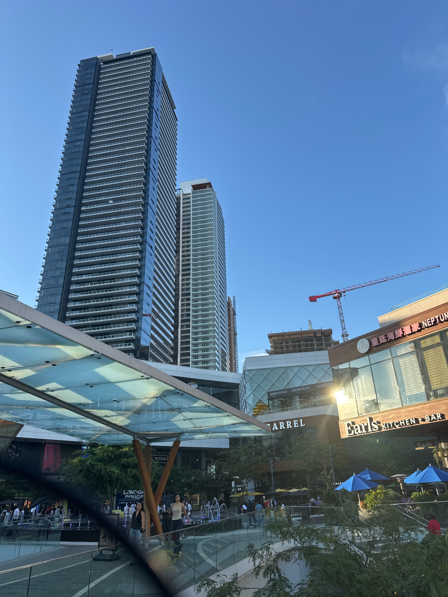 The image depicts a sunny day at a modern urban plaza in Burnaby, Canada, likely Concord Pacific Place, given the architectural style of the buildings. In the foreground, a translucent glass canopy with a metal frame shelters an area with people walking. Beyond the canopy, two tall, sleek residential towers dominate the skyline, their facades characterized by numerous balconies and glass windows. To the right, a commercial building features prominent signage for "Earls Kitchen + Bar" and other businesses, including a Chinese restaurant. The setting sun casts a warm glow, reflecting off the glass surfaces and creating lens flare. Construction is visible in the background with a red crane extending across the sky. Trees and fountains add to the vibrant atmosphere of the public space. People are casually dressed and engaged in various activities, such as walking, dining outdoors under blue umbrellas, and socializing. The overall scene conveys a lively and contemporary urban environment.