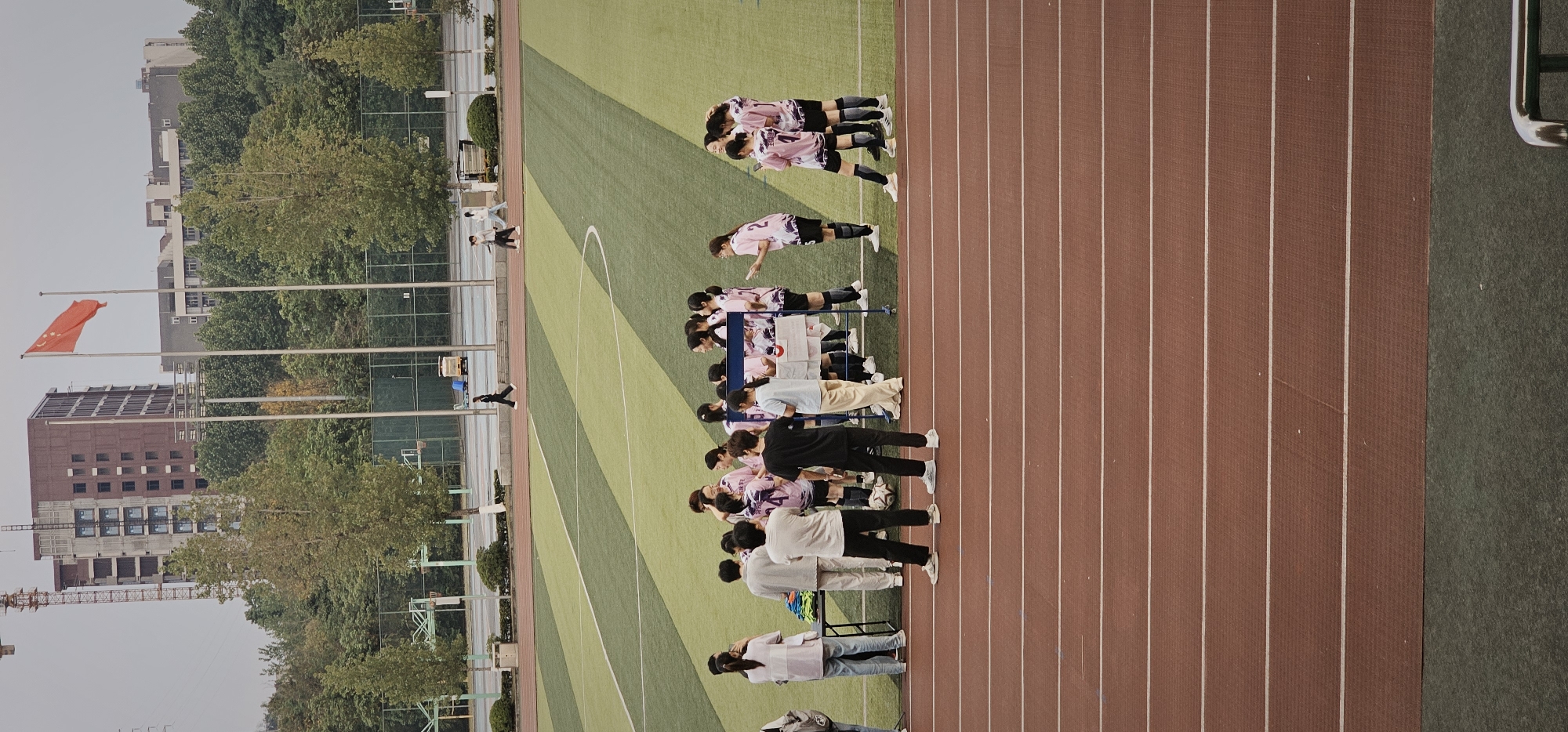 The image captures an outdoor sports complex, likely a university or school campus, situated in the Chongqing High-tech Zone, China. The scene features a vibrant green artificial turf soccer field bordered by a reddish-brown running track.
Numerous young individuals are present, suggesting an organized sports event or practice. On the green field, a group of athletes dressed in matching pink and black athletic uniforms, likely soccer players, are gathered. One cluster appears to be in a huddle, possibly discussing strategy or reacting to a play, while another individual in a similar uniform is seen bending over.
Along the running track adjacent to the field, a larger group of people is observing the activities. Some are also in the pink and black uniforms, while others wear casual attire such as t-shirts, hoodies, and jeans, indicating a mix of participants and spectators. A soccer ball rests near this group, and a few benches or tables are set up for their use. Further back on the track and near the field's sidelines, other individuals are walking or standing.
In the background, behind the sports field, multiple multi-story buildings are visible, interspersed with abundant green trees. One building prominently features elements of ongoing construction, with a visible crane nearby. Several flagpoles stand tall, one proudly flying a red Chinese flag, confirming the geographical location. The sky is overcast or hazy, suggesting daytime conditions with diffused natural light. No discernible text is visible on uniforms or any signage.