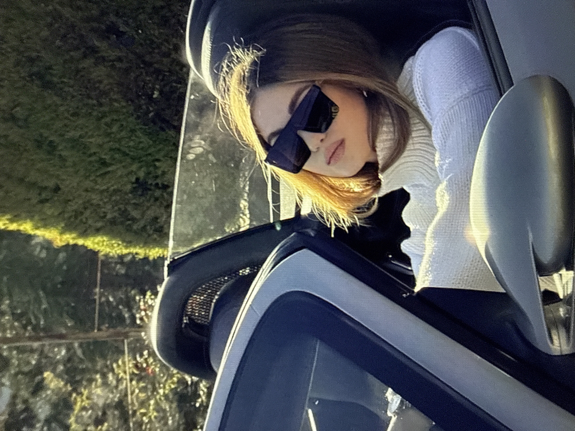 The scene depicts a person seated in the driver's seat of a convertible vehicle, observed outdoors under bright sunlight. The location is stated to be in the city of Kardzhali, Bulgaria.
The primary subject is an individual, appearing to be female, with medium-length, light brown hair significantly highlighted by direct sunlight. She is wearing large, dark, rectangular-framed sunglasses that obscure most of her eyes. Her lips are visible and appear to have a neutral or slightly parted expression. The individual is dressed in a white, ribbed turtleneck sweater, and her arms are crossed at the chest. Her head is tilted slightly upward and to her left, indicating a gaze directed away from the camera and towards the sun.
The vehicle is a light metallic grey or silver convertible sports car, evidenced by the visible driver's side A-pillar, windshield frame, and a dark-colored roll-hoop or wind deflector system positioned behind the seats, featuring a mesh insert. A portion of the dashboard and the driver's side mirror are also visible in the foreground.
The background consists primarily of out-of-focus green foliage, including trees and dense shrubs, suggesting a natural or landscaped outdoor environment. A faint vertical linear element, possibly a utility pole or fence post, is discernible in the upper left background. The lighting is characterized by strong, direct sunlight, creating bright highlights on the subject's hair and sweater and illuminating the scene from the upper left.