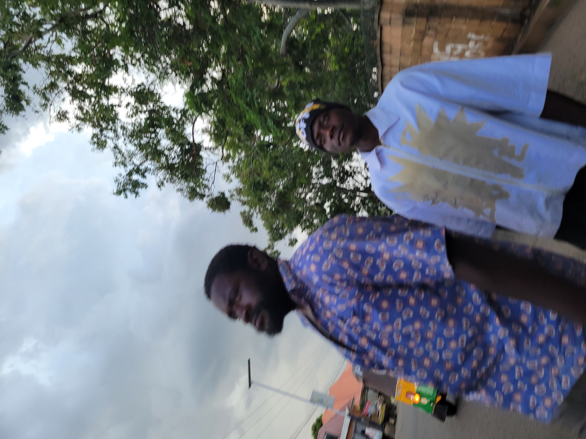 Two men are pictured on a street in Jos, Nigeria, with a cloudy sky overhead. The man in the foreground on the left is wearing a blue patterned shirt and looking upwards and to the left. He has dark skin, a beard, and appears to be in motion. To his right and slightly behind him, a second man is wearing a white zippered shirt and a black beanie with yellow and white designs. He is looking forward. Behind the men, a brick wall runs along the side of the street, and a green and yellow motorized rickshaw is visible further down the road. Trees with lush green foliage are also present in the background. The lighting suggests it is either early morning or late afternoon.