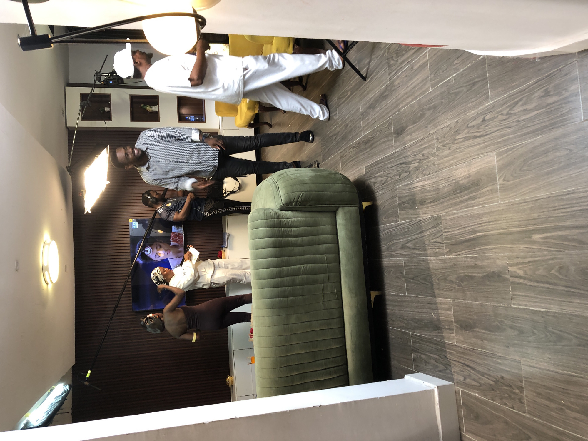 This image captures an indoor video production setup within a modern space. The floor features gray wood-look tiles, and a dark brown, ribbed accent wall serves as a backdrop for a large flat-screen television. An olive green, ribbed sofa is prominently placed in the foreground.
Multiple individuals are engaged in the filming process. On the right, a person dressed in all white holds a large lighting diffuser, while another person in a striped shirt stands nearby, smiling, with a rectangular softbox light illuminating the area. Behind them, a crew member wearing headphones appears to be monitoring audio or camera operations.
In front of the television, which displays a close-up of a woman's face (with faint "KLG" text visible in the top right), a person in a white outfit and head wrap holds a script or document, seemingly the subject of the filming. Another person in a dark, form-fitting outfit and head wrap observes the activity from the left. A camera on a tripod is positioned to capture the scene, and a boom microphone extends overhead to record audio. Additional lighting fixtures are strategically placed to ensure proper illumination. The overall scene suggests a professional and focused media production.