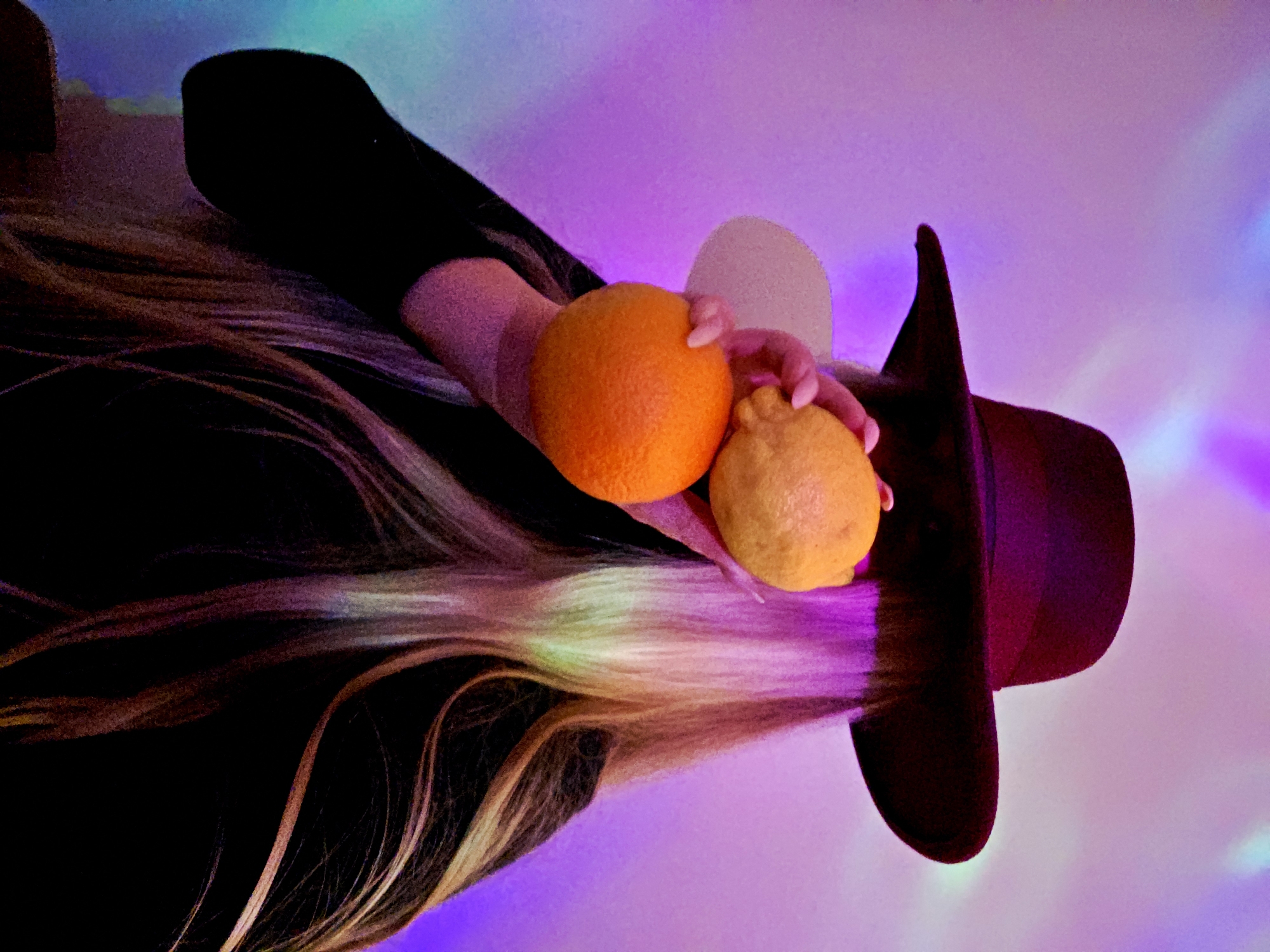 The image displays a person's upper body, with a focus on their hands, hair, and a hat, set against a vibrantly lit background. The perspective is from above and to the side, with the image appearing rotated, making the person's head and hat on the right and their hair flowing to the left.
The main subject is a person, whose face is not visible. They are wearing a dark, possibly black, long-sleeved garment and a dark hat, which appears to be a fedora or similar style with a soft brim. Their long, light-colored hair, potentially blonde or light brown, flows down the left side of the frame, catching the colorful ambient light and showing streaks of pink and purple.
The person is holding two pieces of fruit: a bright orange in their left hand (closer to the viewer) and a yellow lemon in their right hand. The hands appear to be gently grasping the fruits.
The background is abstract and softly blurred, composed of swirling and blending colors, predominantly vivid purples and pinks, with accents of blue and green light. This suggests an indoor setting with atmospheric or mood lighting, possibly from LED lights or a disco ball, creating a dreamlike or artistic ambiance.
No specific activity is taking place beyond holding the fruits. The lighting indicates it is likely evening or night. There are no discernible emotions due to the absence of facial features. No text is visible in the media file.