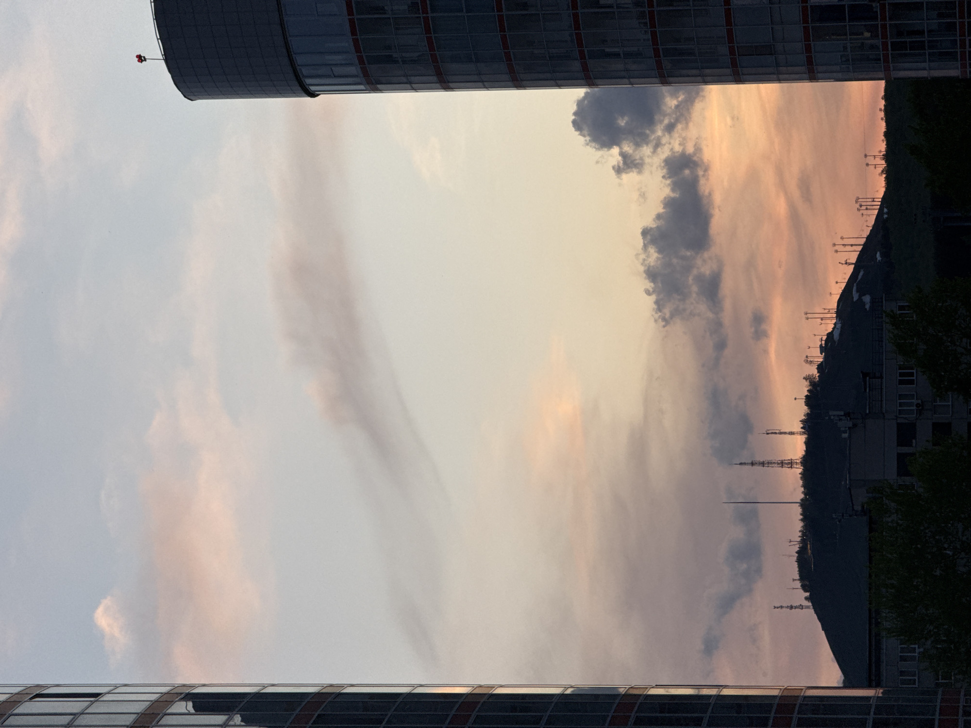 The image, shot from an unusual angle, captures the sky framed by parts of buildings. The sky is a blend of pale blues, whites, and hints of pink and peach, suggesting either sunrise or sunset. There are some scattered, dark clouds. In the background, a hill can be seen, dotted with multiple communication towers. There is some residential buildings and greenery in the foreground, partially obscured by the angle and lighting. From context clues, the location is Krasnoyarsk, Russia.