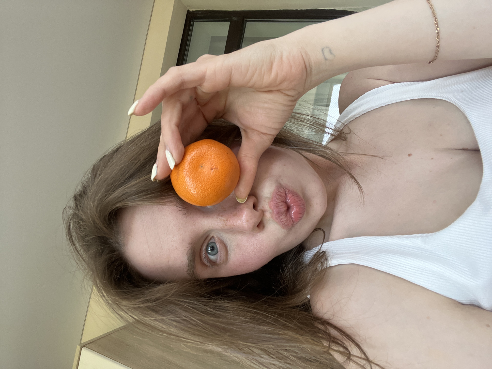 The scene is situated in the city of Chaikovsky, Russia. The image displays a young woman in a close-up, head-and-shoulders shot, facing the camera directly. She has light brown hair, fair skin with visible freckles across her nose and cheeks, and her left eye, which is blue, is wide open. Her lips are pursed in a "kissy" expression. She holds a small, bright orange mandarin with her right hand, positioning it to cover her right eye. Her fingernails are long, almond-shaped, and painted white. She is wearing a white ribbed tank top. A thin, gold-toned chain bracelet is present on her right wrist, along with a small, faint, heart-shaped tattoo on the inner side of the same wrist. The background includes a light-colored ceiling, a partial view of a light wooden cabinet or structure on the upper left, and a dark-framed window or glass door on the right.