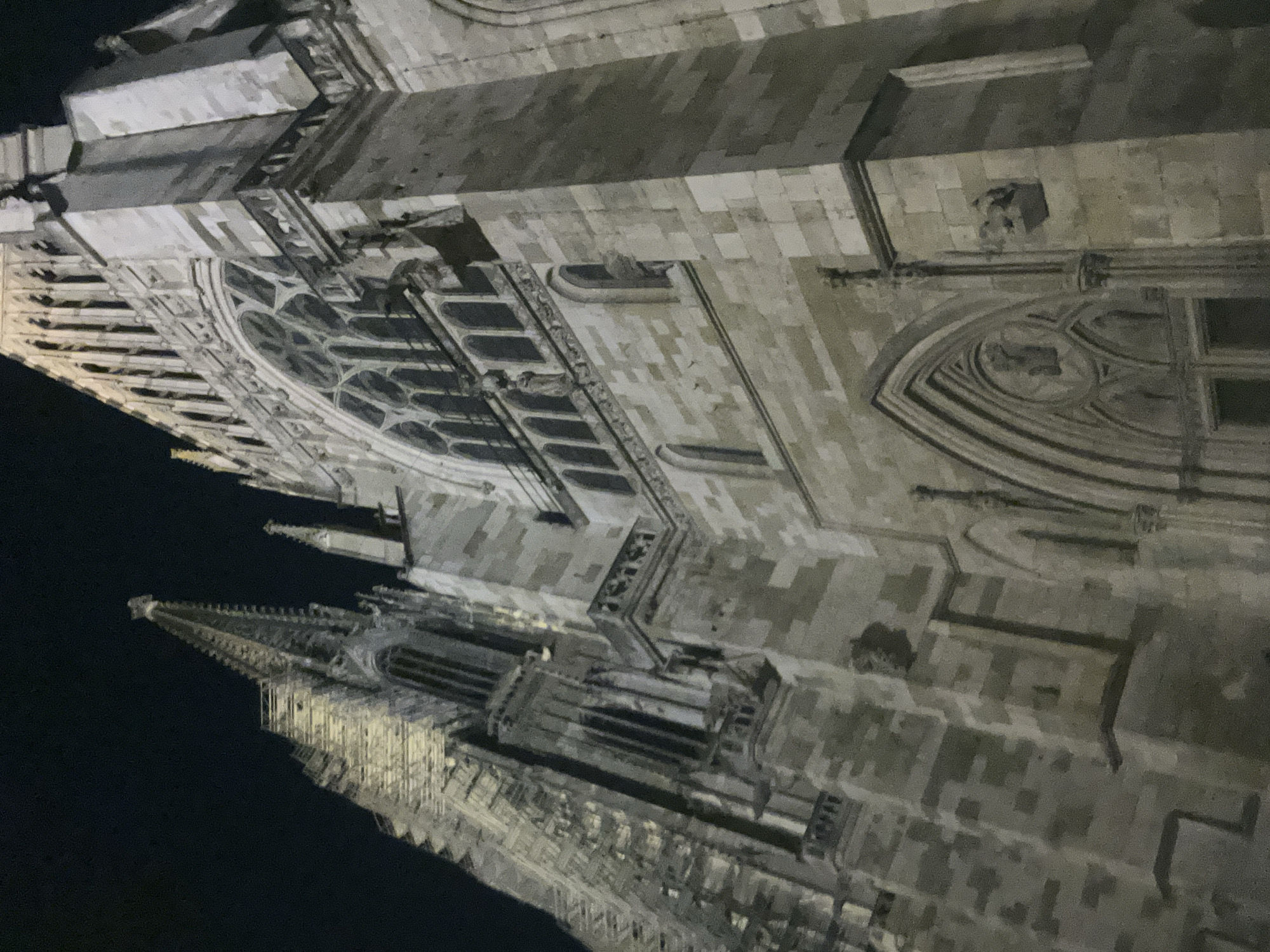 The image shows a nighttime view of the Regensburg Cathedral (Dom St. Peter) in Regensburg, Germany. The camera is angled upwards, capturing the imposing Gothic architecture of the cathedral's facade and spires. The stone structure is illuminated, creating strong contrasts between light and shadow. Several architectural details are visible, including pointed arches, intricate carvings, and tall, narrow windows. The dark night sky provides a stark backdrop to the illuminated building.