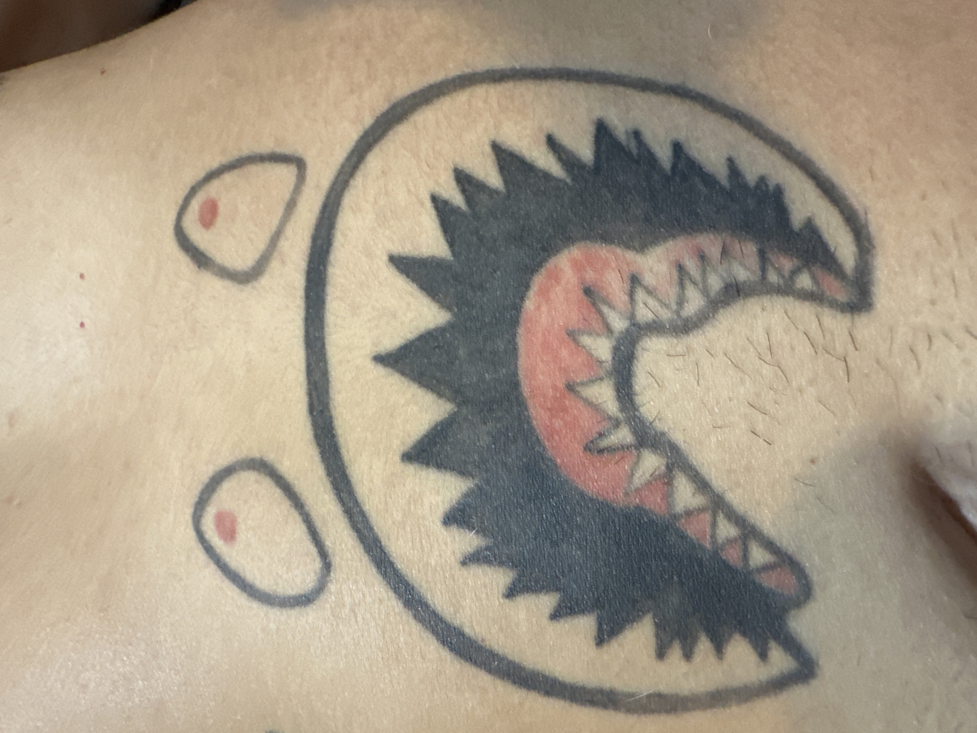 A close-up shot reveals a tattoo on human skin. The tattoo design is a stylized Pac-Man character, but with a shark-like mouth filled with jagged teeth and a red inner lining. Two additional, smaller Pac-Man eye designs, each with a red pupil, are positioned above and to the left of the main figure. The skin tone appears light. No context suggests Buenos Aires, Argentina.