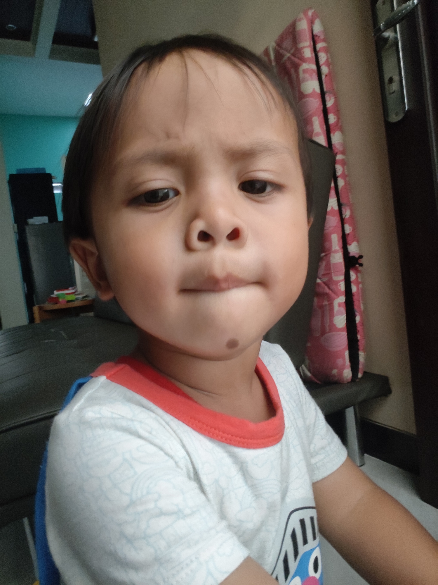 A young child, appearing to be of Southeast Asian descent, is pictured in a close-up portrait. The child has dark hair, dark eyes, and fair skin with a slightly rosy complexion. A small, dark mole is visible on the right side of their chin. The child is wearing a white t-shirt with a red neckline and a blue sleeve. The background is softly blurred, showing a teal wall to the left and a door with a metallic handle to the right. A pink and white patterned object, possibly a cushion or bag, is leaning against a dark chair in the mid-ground. A shelf with some items is also visible behind the child. The overall lighting suggests an indoor setting. The image is a still photograph. Without further information or visual cues, it is not possible to determine the specific location within Pemalang, Indonesia.