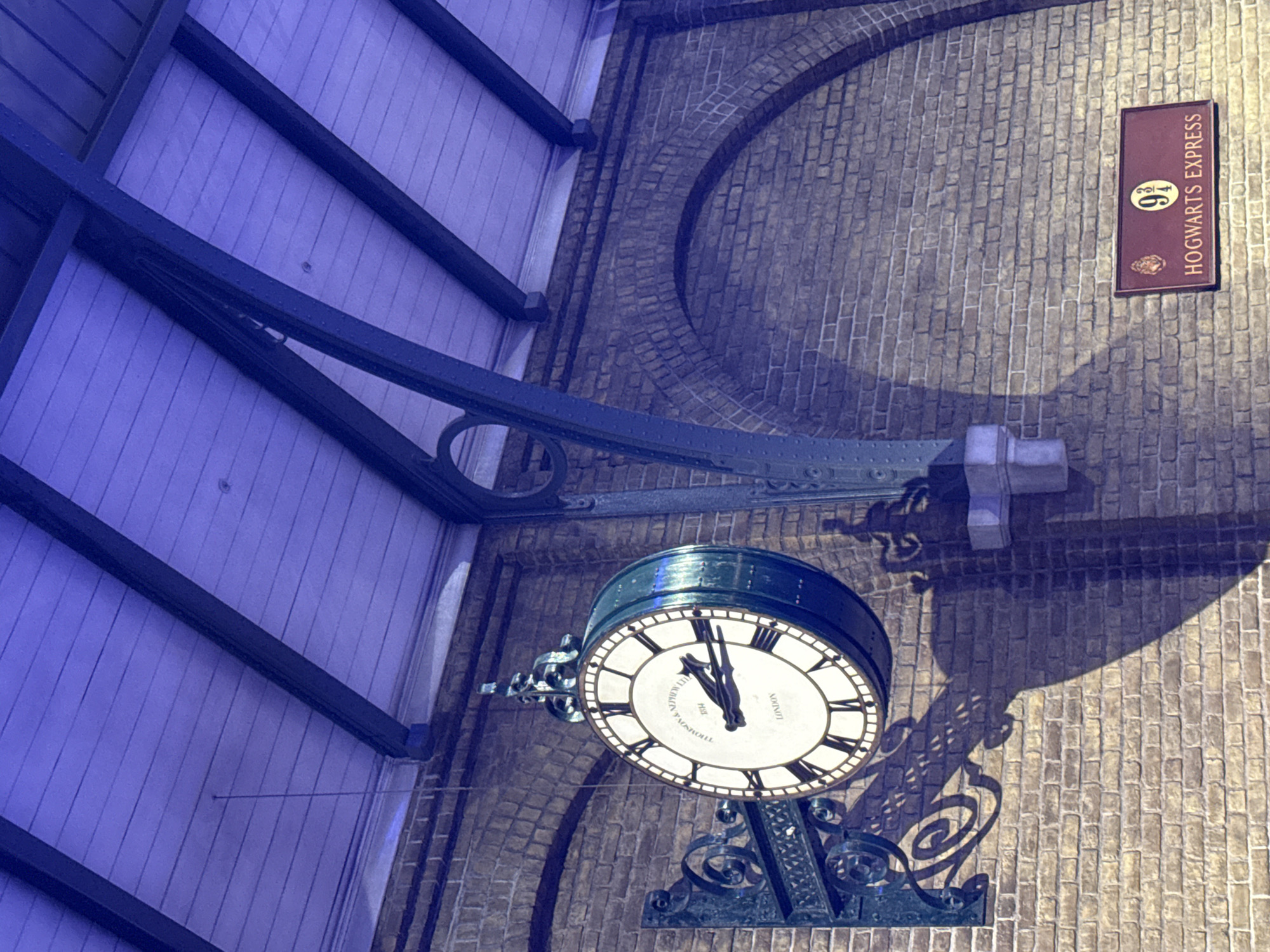 The image shows a brick wall with a vintage clock hanging on it. The clock is round with Roman numerals and is supported by an ornate metal bracket. Above the clock, a wooden ceiling with exposed beams is visible, illuminated by a purple light. To the right of the clock, a sign with "9 3/4" and "HOGWARTS EXPRESS" is mounted on the brick wall. The scene appears to be an indoor setting, possibly a themed attraction or a film set. The lighting creates a dramatic effect with strong shadows cast by the clock and its bracket on the wall. The location is specified as the city of Three Rivers, United Kingdom.