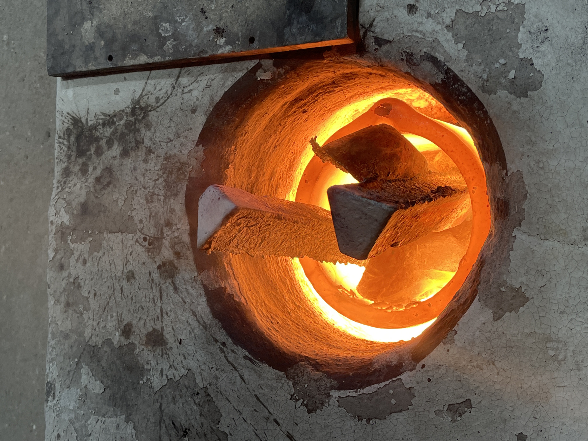 The image depicts a furnace with a circular opening, revealing a bright orange interior. Within the furnace, there are two pieces of metal, possibly triangular or rectangular in cross-section, positioned diagonally across the opening. The furnace's exterior wall is textured and appears to be made of concrete or a similar material, with a surface that is cracked and peeling. The location, Saint-Rémy-lès-Chevreuse, France, is not directly discernible from the visual elements of the image itself.