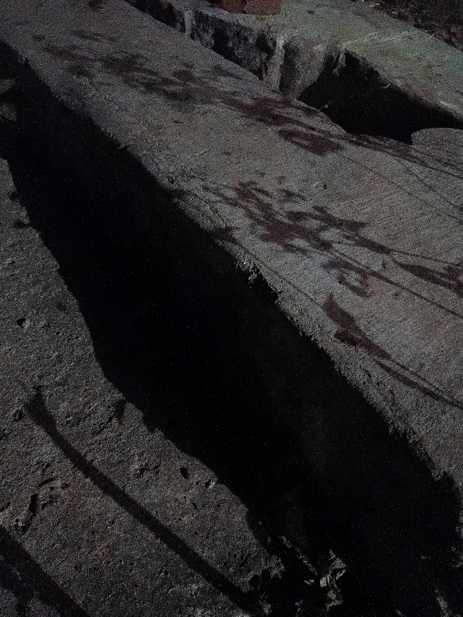 The image depicts a section of a dark, rough-textured surface, possibly concrete or asphalt, with a deep, shadowed crevice running diagonally across it. The crevice is so dark that details within it are obscured. On the illuminated parts of the surface, there are faint, mottled shadows that suggest the presence of overhead foliage or structures casting them. The overall impression is one of a gritty, urban environment, perhaps an alleyway or a less-maintained walkway. There are no discernible people or specific objects that would allow for precise identification of a location within Cikal, Indonesia. The scene is devoid of color, rendered in shades of gray and black, with a grainy texture throughout.
